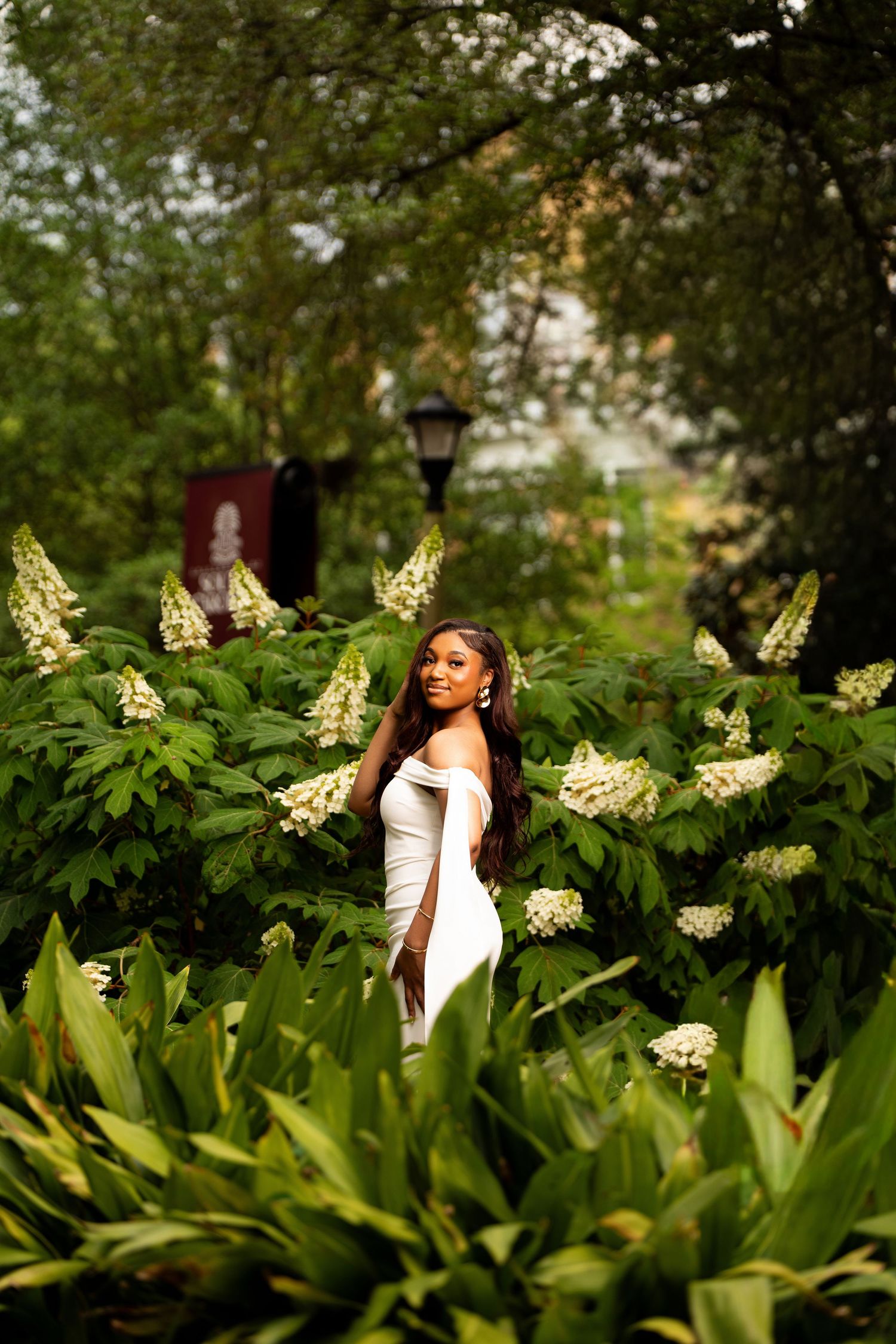 A woman in a white off-shoulder dress poses among lush green foliage and white hydrangea blooms in a garden.