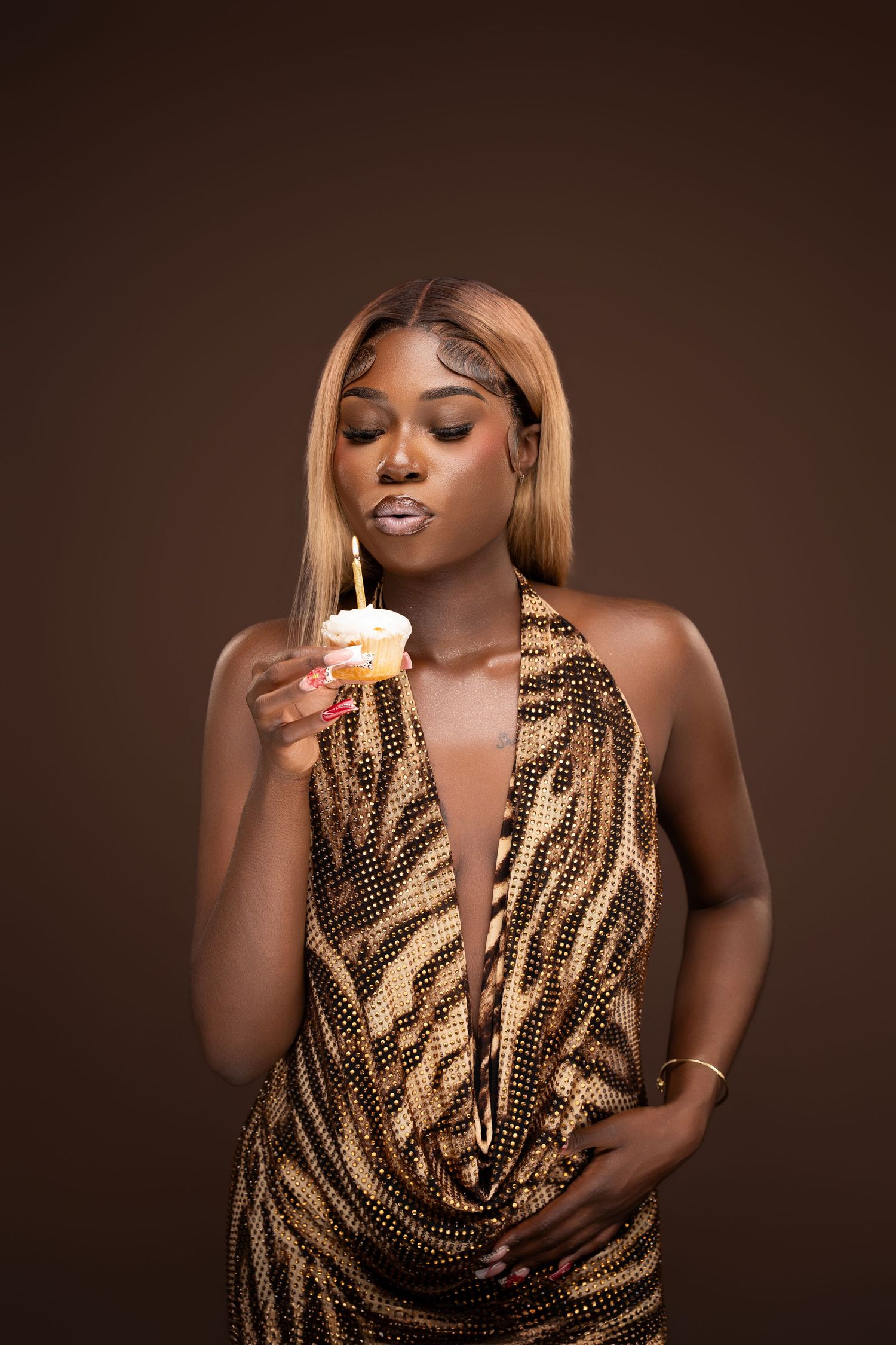 Elegant Black woman in gold sequin dress blowing out birthday candle on cupcake against brown background.