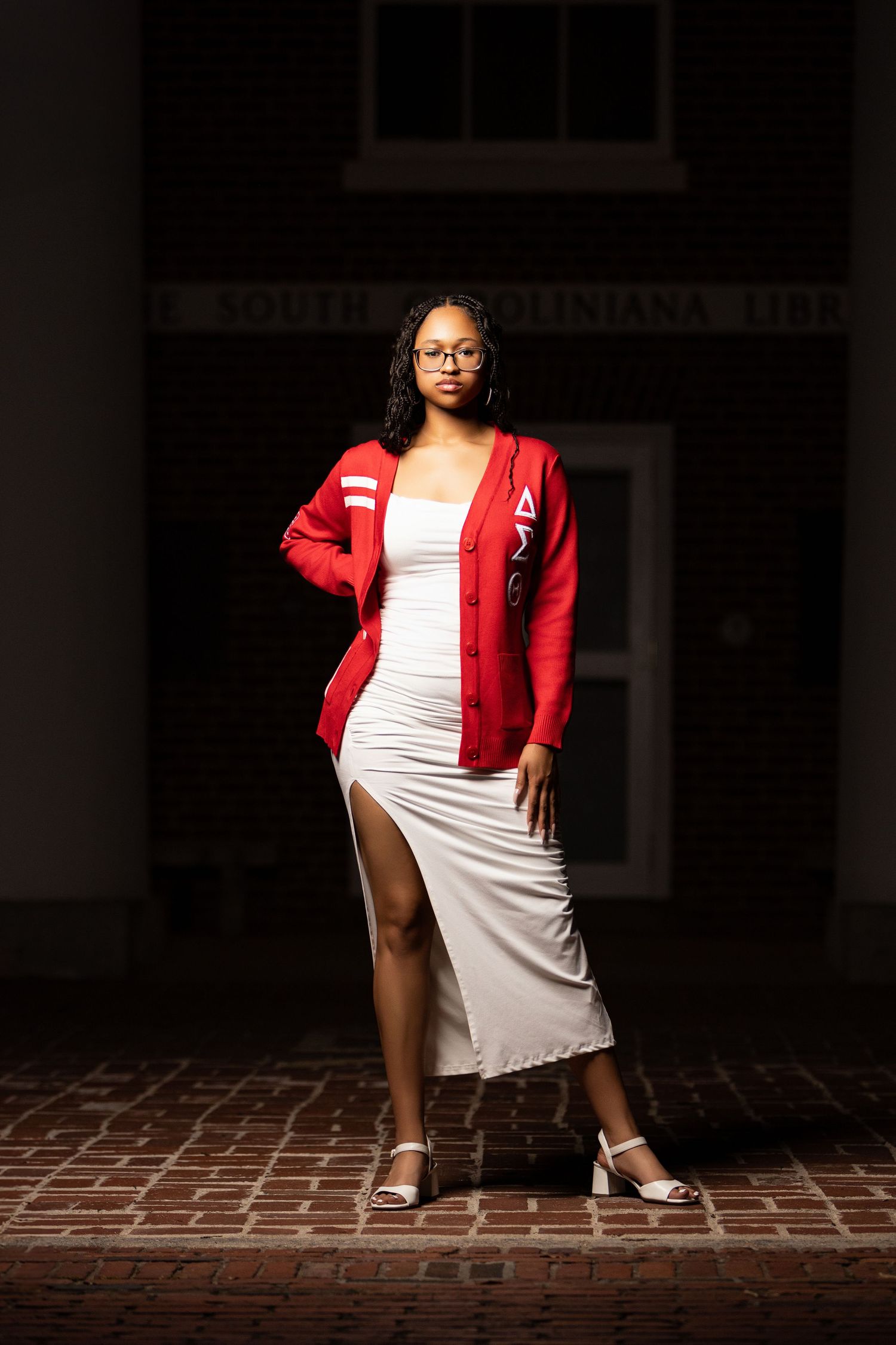 Street fashion pose with red letterman jacket and white dress against brick background.