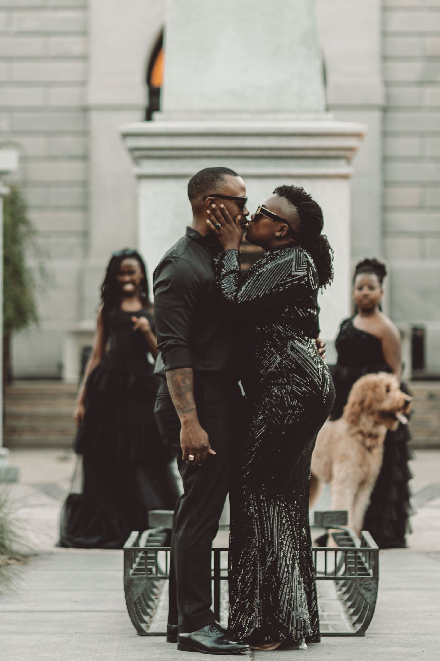 Couple embraces during formal event with wedding party and dog in background.