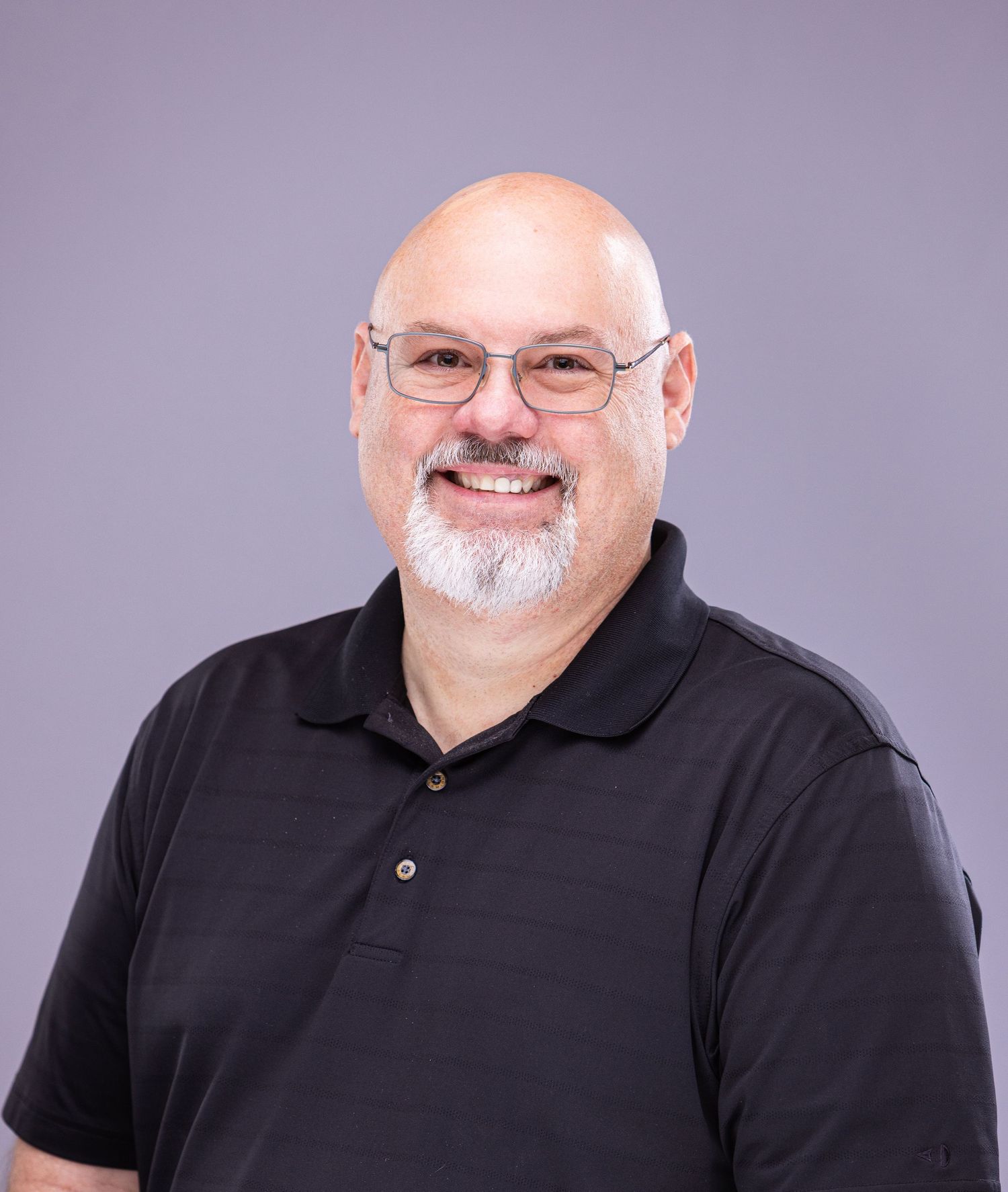 Professional headshot of person in black polo shirt with glasses and white beard against purple backdrop.