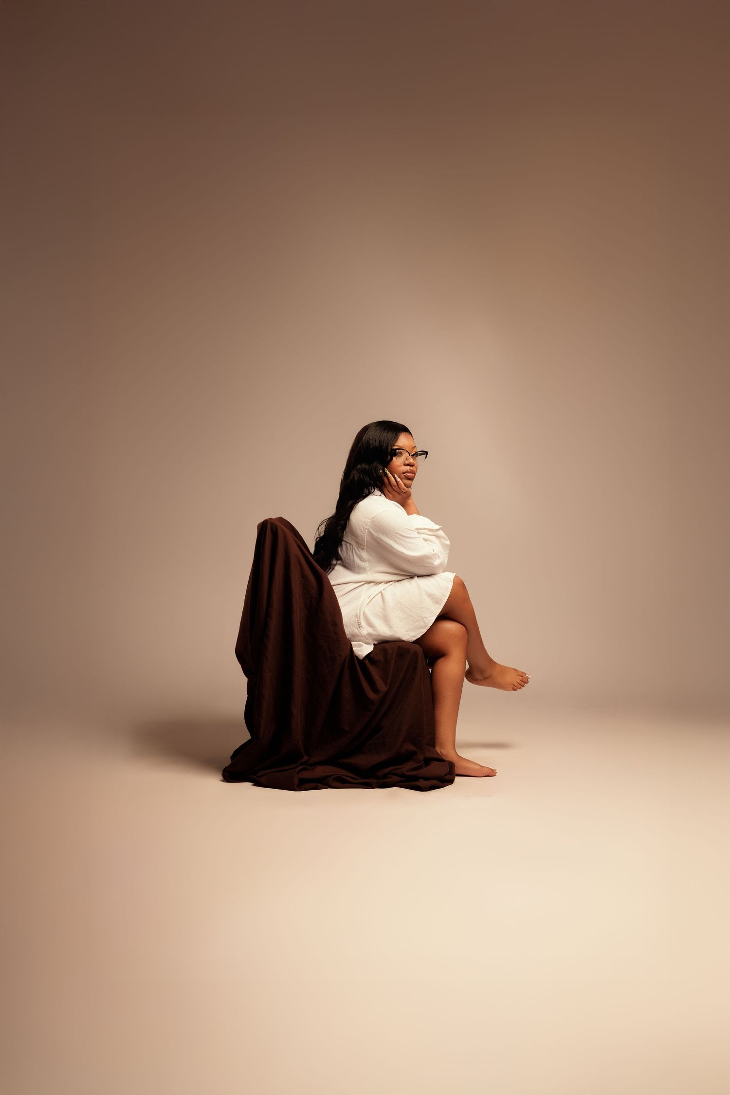 A person in a white top sits cross-legged on a brown fabric against a neutral studio background.