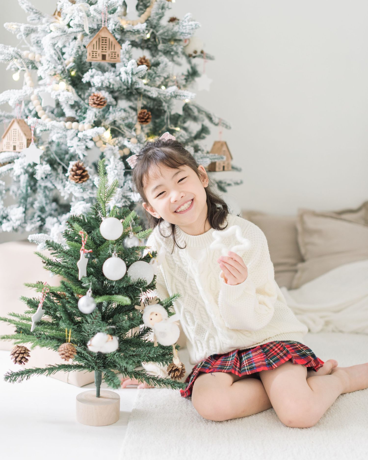 Child in cream sweater and plaid skirt poses by decorated Christmas tree with white ornaments and pinecones.