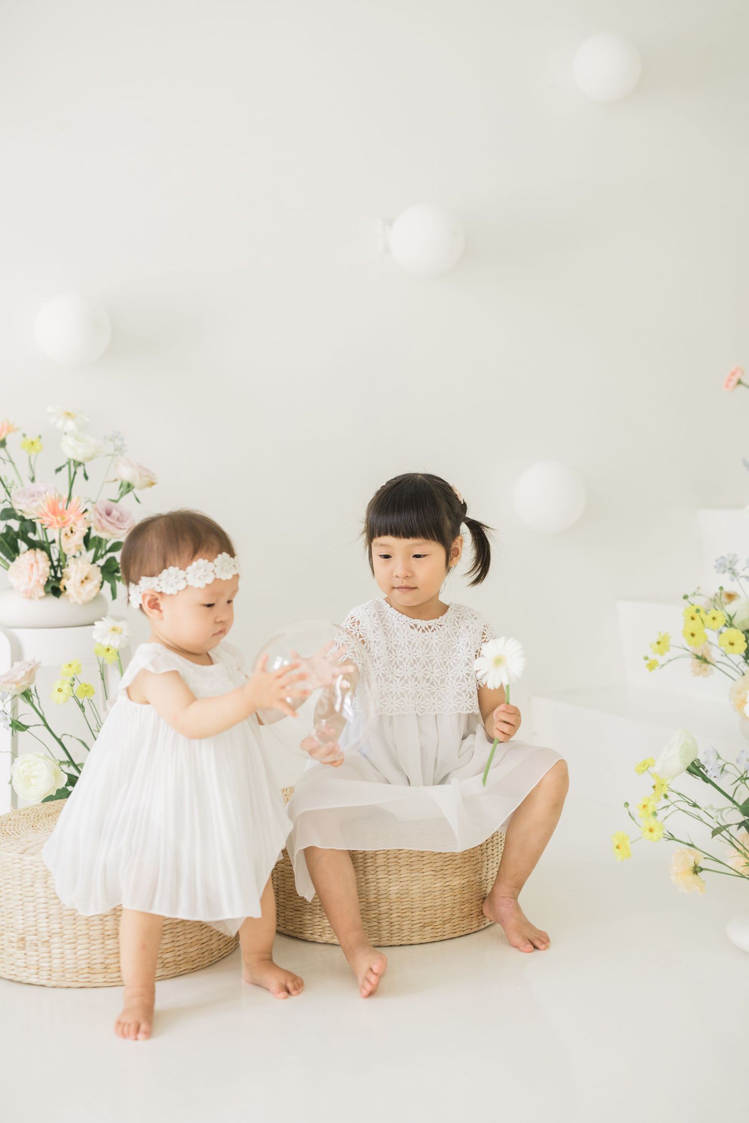 Two young girls in white dresses sit among flowers and balloons in a bright, airy studio photo session.