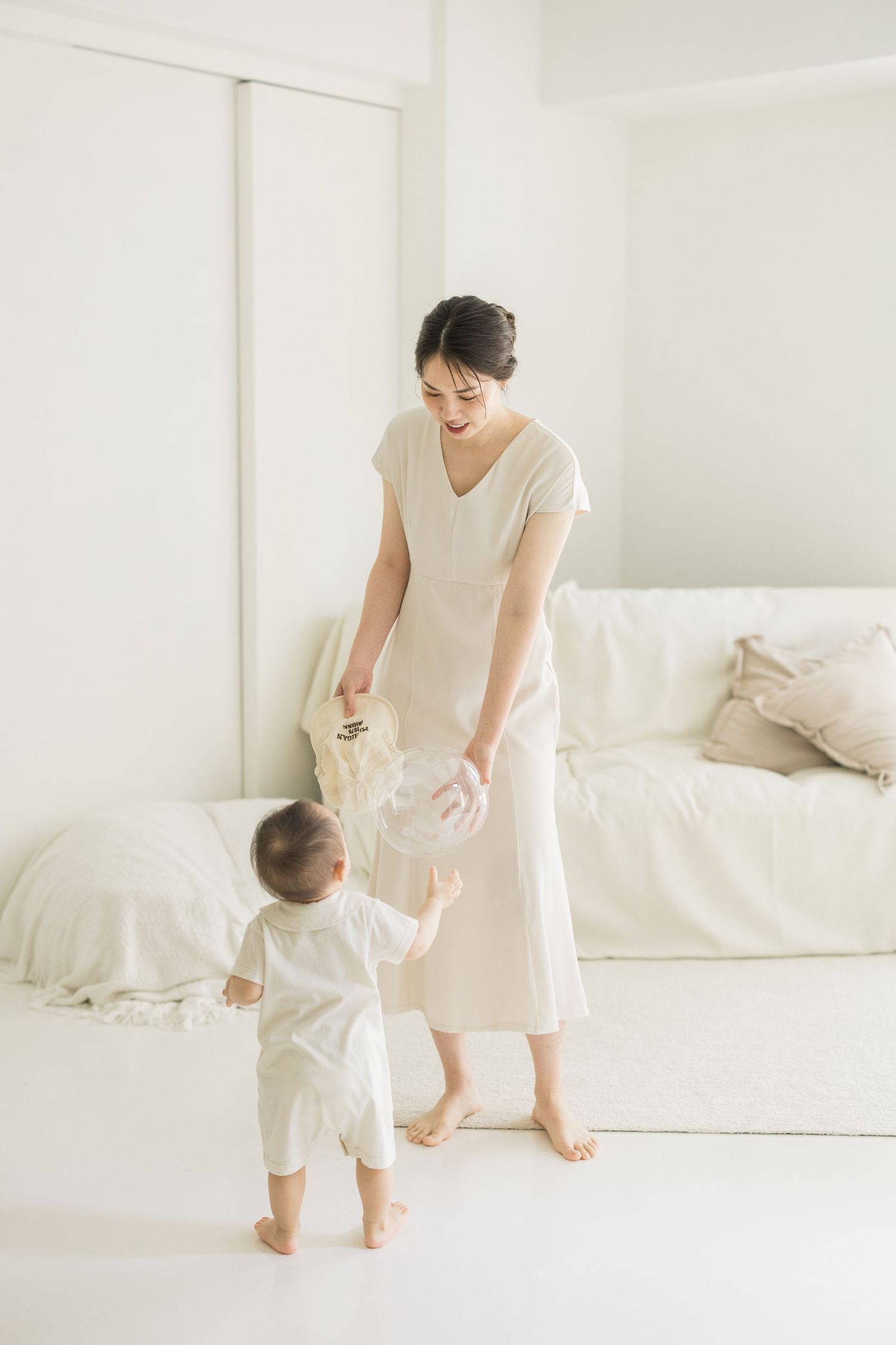 A young Asian mother in a beige dress plays with her toddler in a bright white minimalist living room.