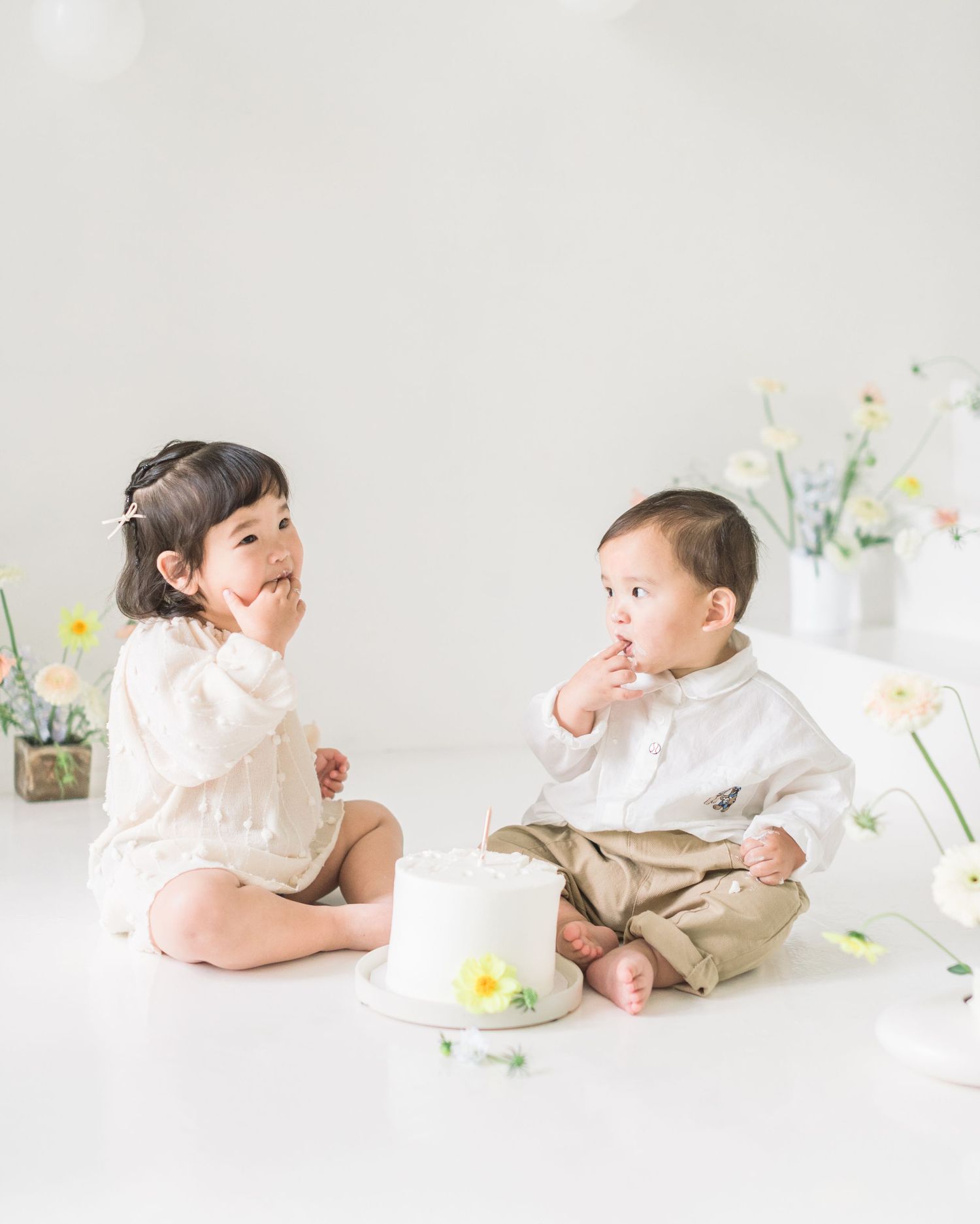 Two small children in light colored clothing sit on a white floor with delicate floral decorations.