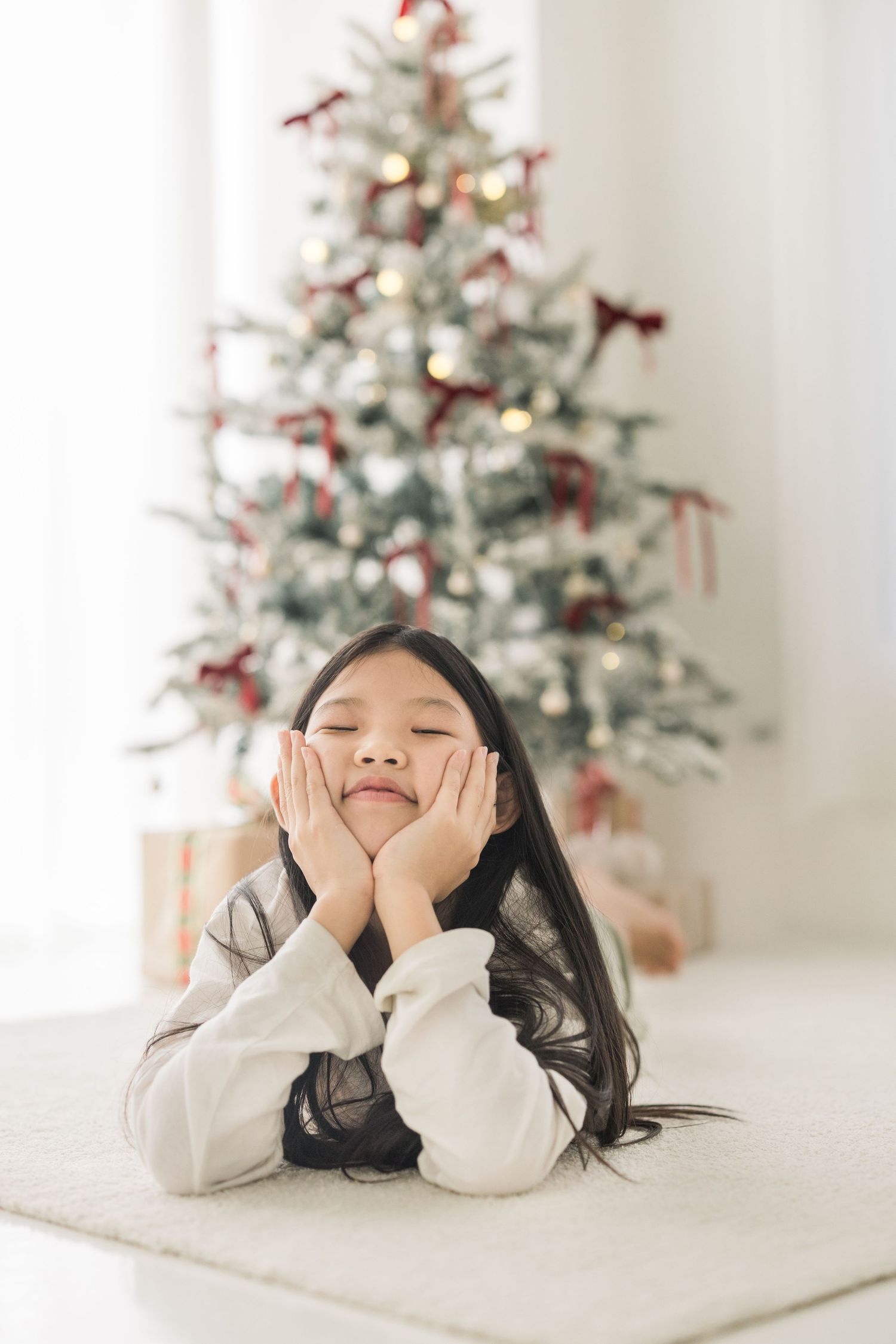 A child lies on a white floor making playful expressions with a decorated Christmas tree in the background.