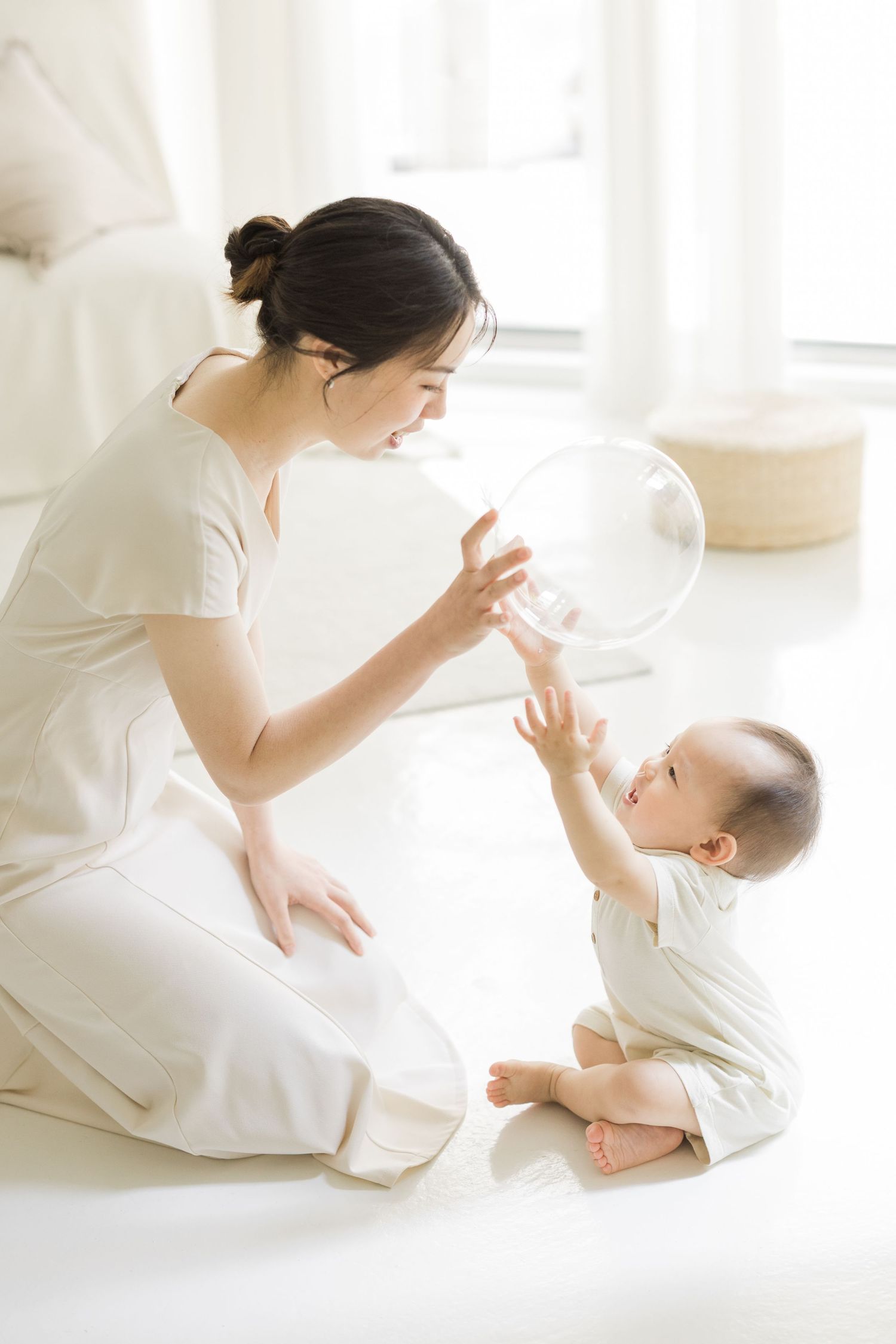 A young Asian mother in white dress feeds her baby with a bottle while sitting on a bright white floor.