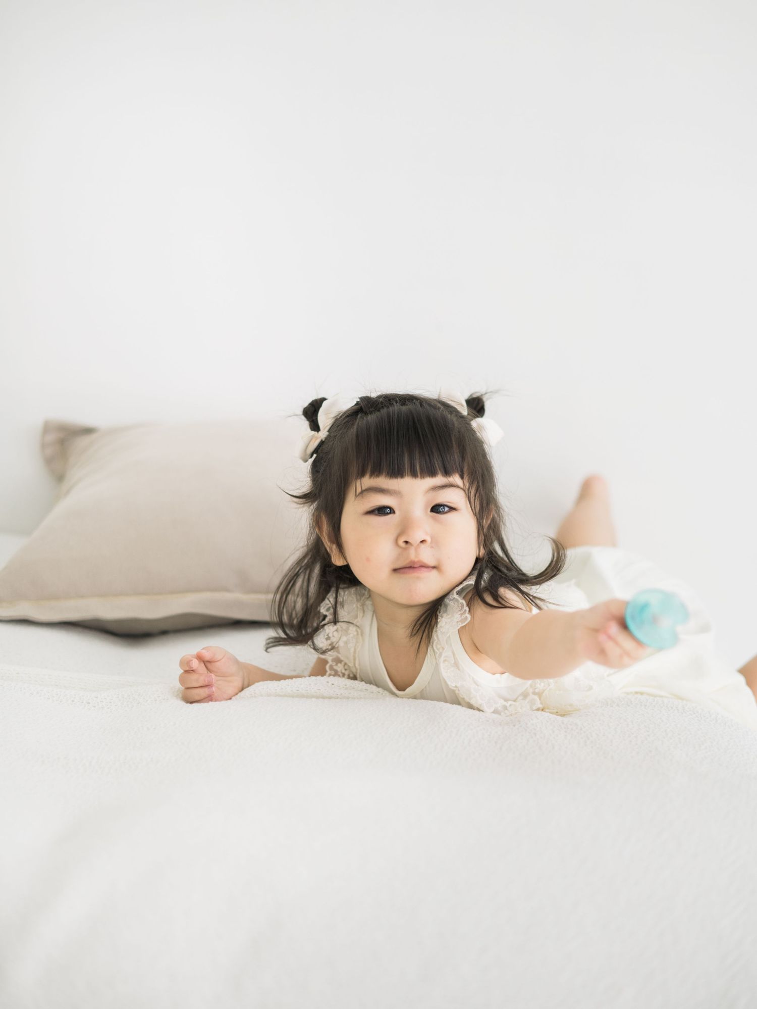 A young child in white lies playfully on soft white bedding in a bright, minimalist setting.