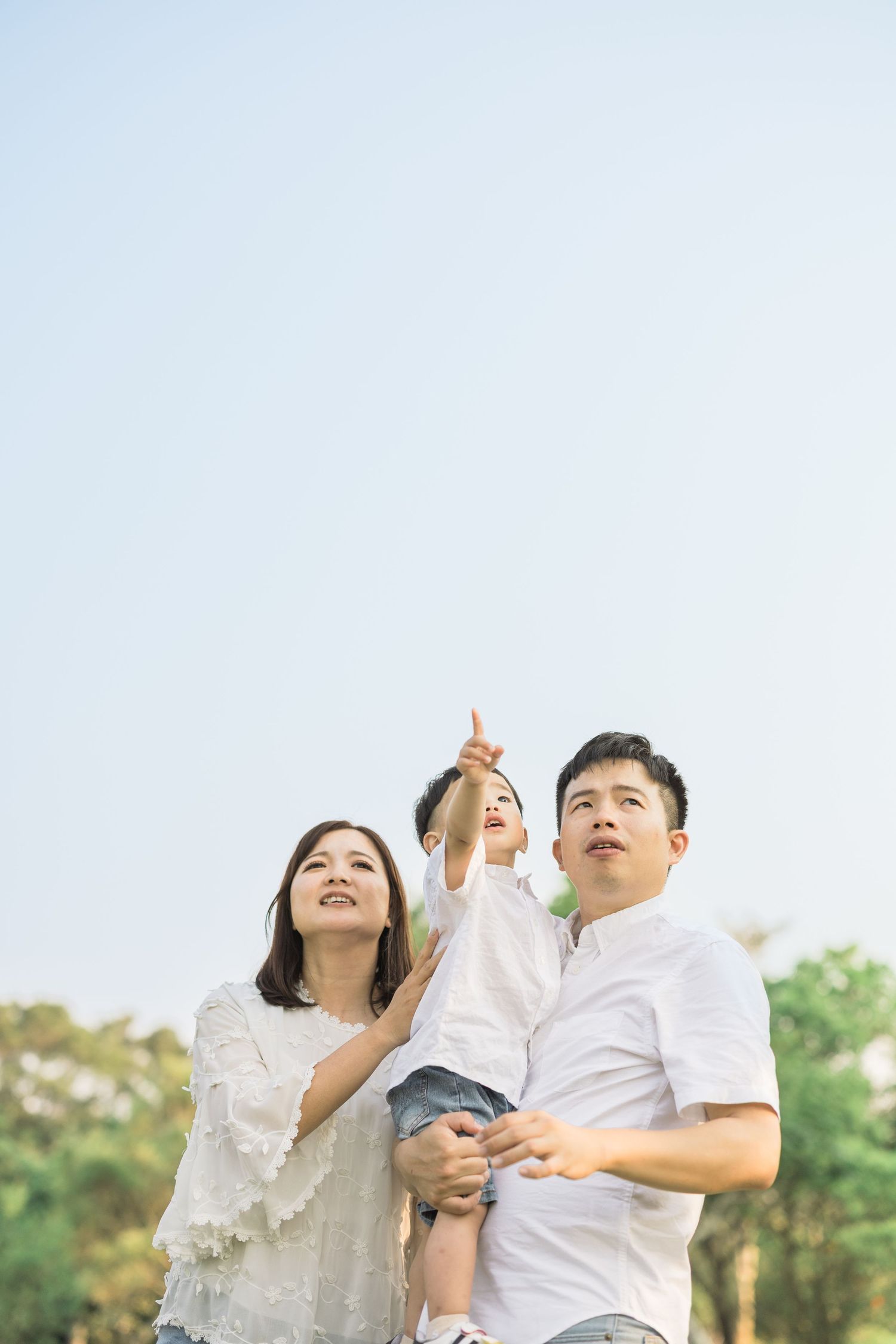 Happy Asian family with parents holding toddler pointing at sky in sunny outdoor park setting.