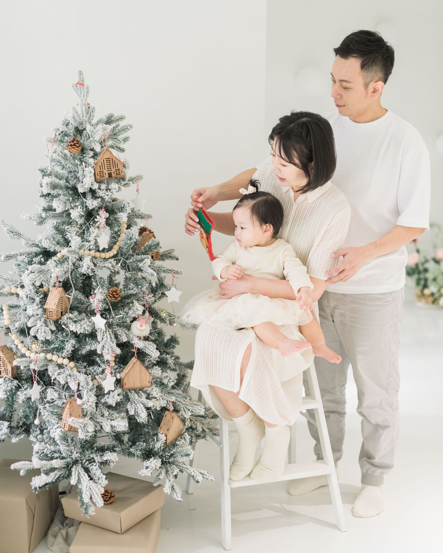 A parent and child decorate a flocked Christmas tree together in a bright white room with soft natural lighting.
