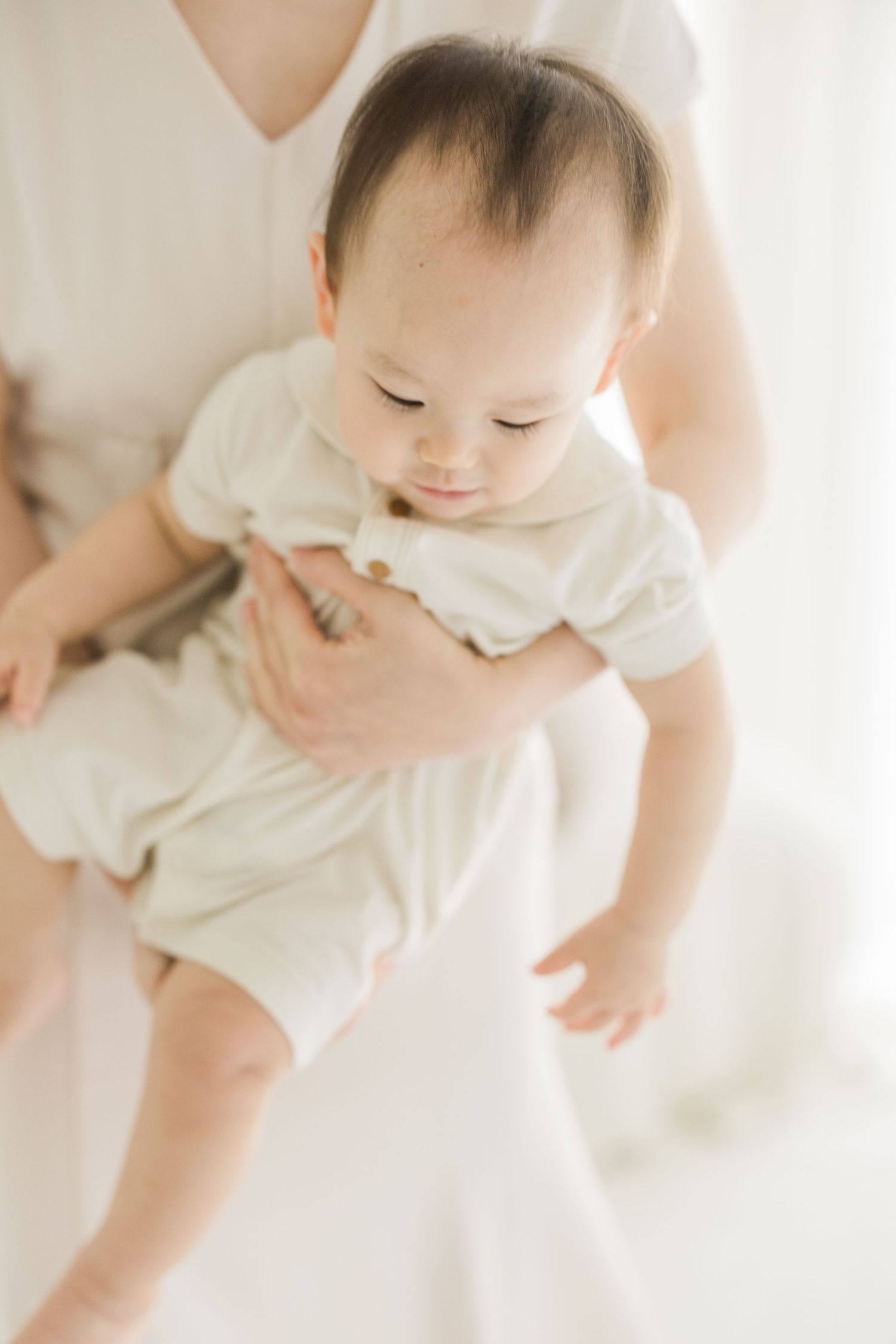 A mother in white dress gently holds her smiling baby in a cream onesie in a bright, airy room.