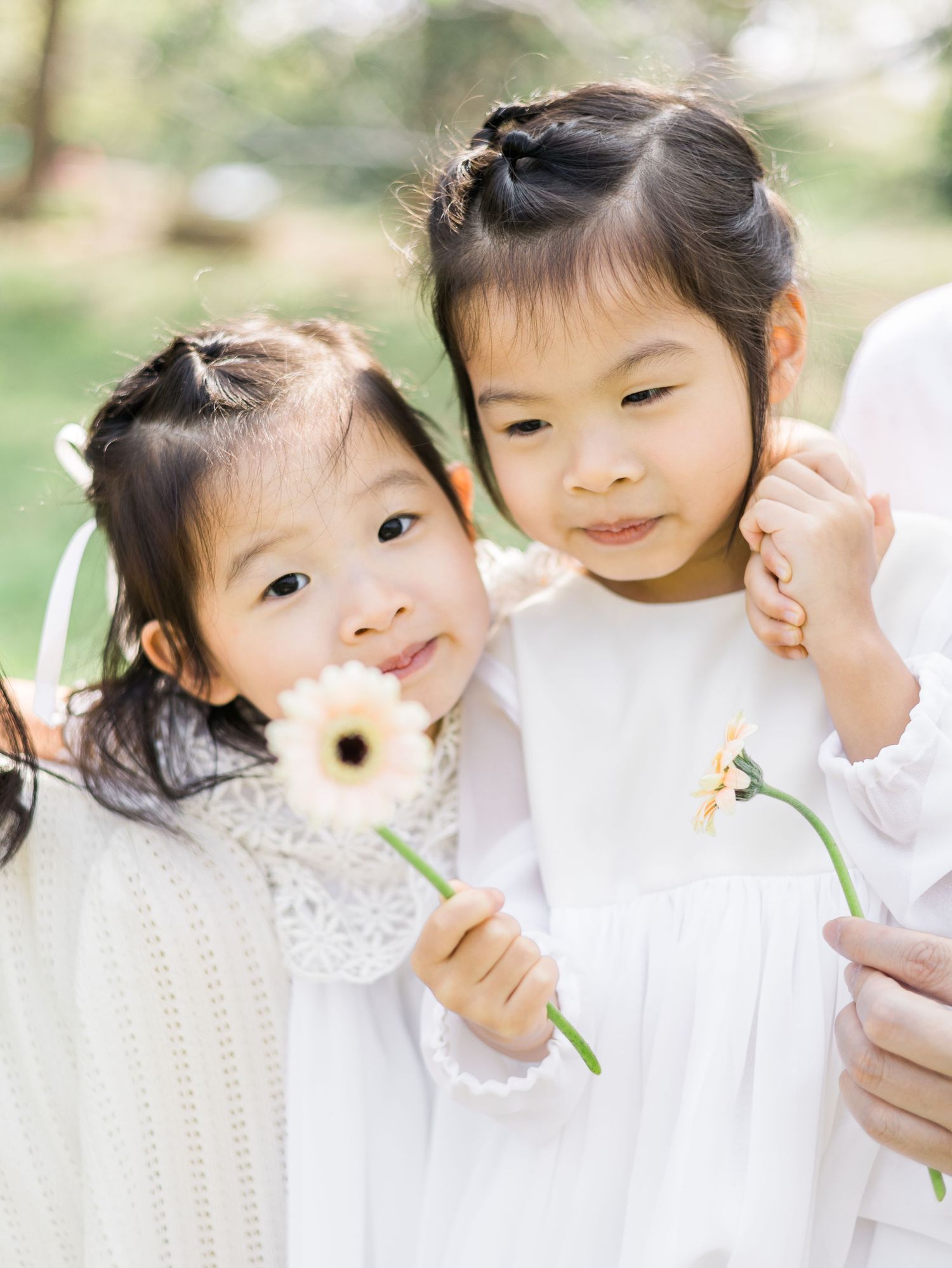 Two children in white dresses holding flowers while posing together in a garden.
