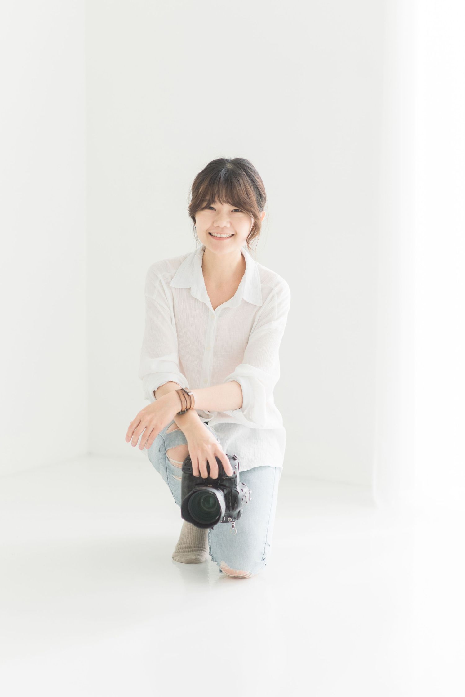 A photographer in a white shirt and jeans kneels with a camera in a bright white studio setting.