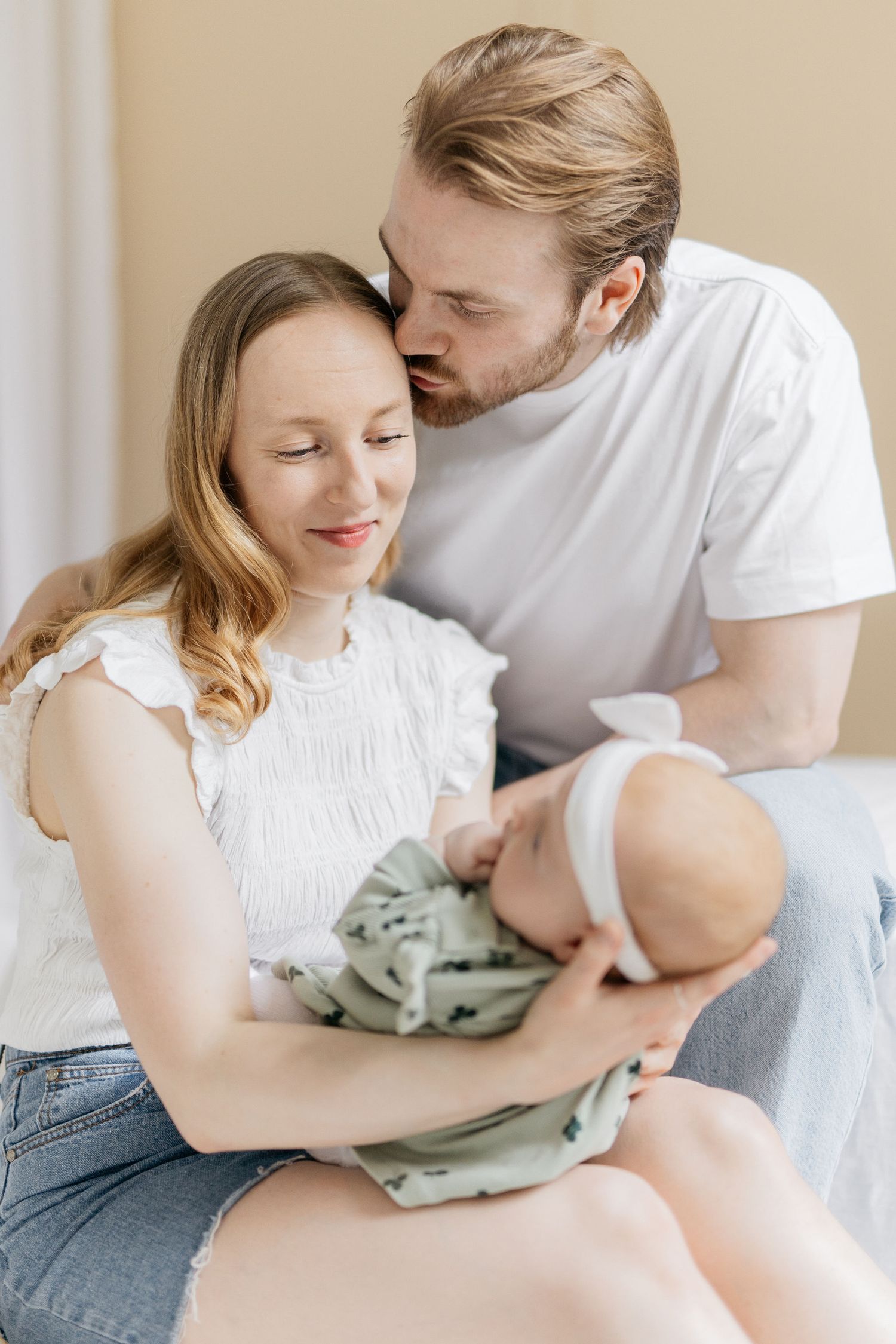 Perhekuvaus seinäjoella. Äiti katsoo vauvaa ja isä suutlee kevyesti äitiä ohimolle