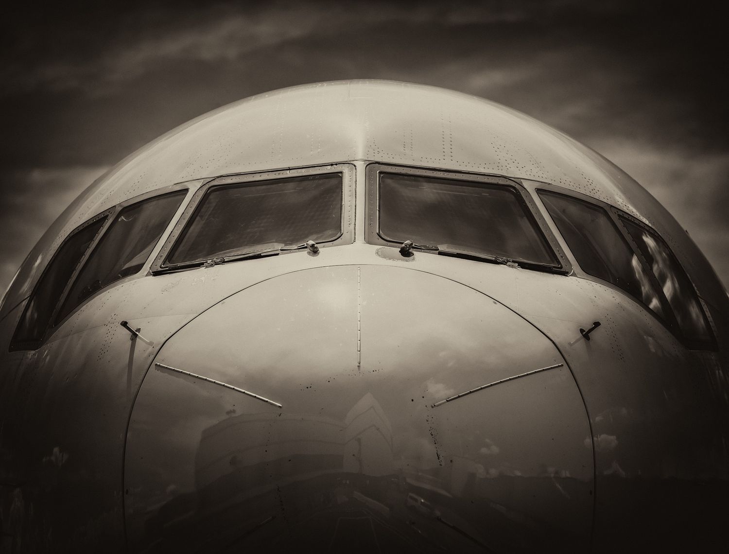 Sepia close-up of a commercial airliner's nose and cockpit windows under a moody, cloudy sky.