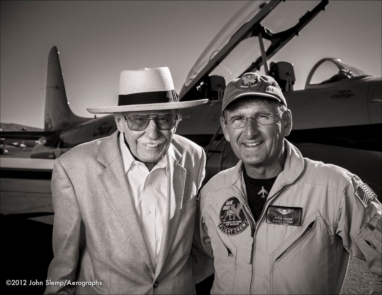 Two smiling men pose in front of a jet aircraft, one in a white hat and sport coat, the other in a flight suit and cap.