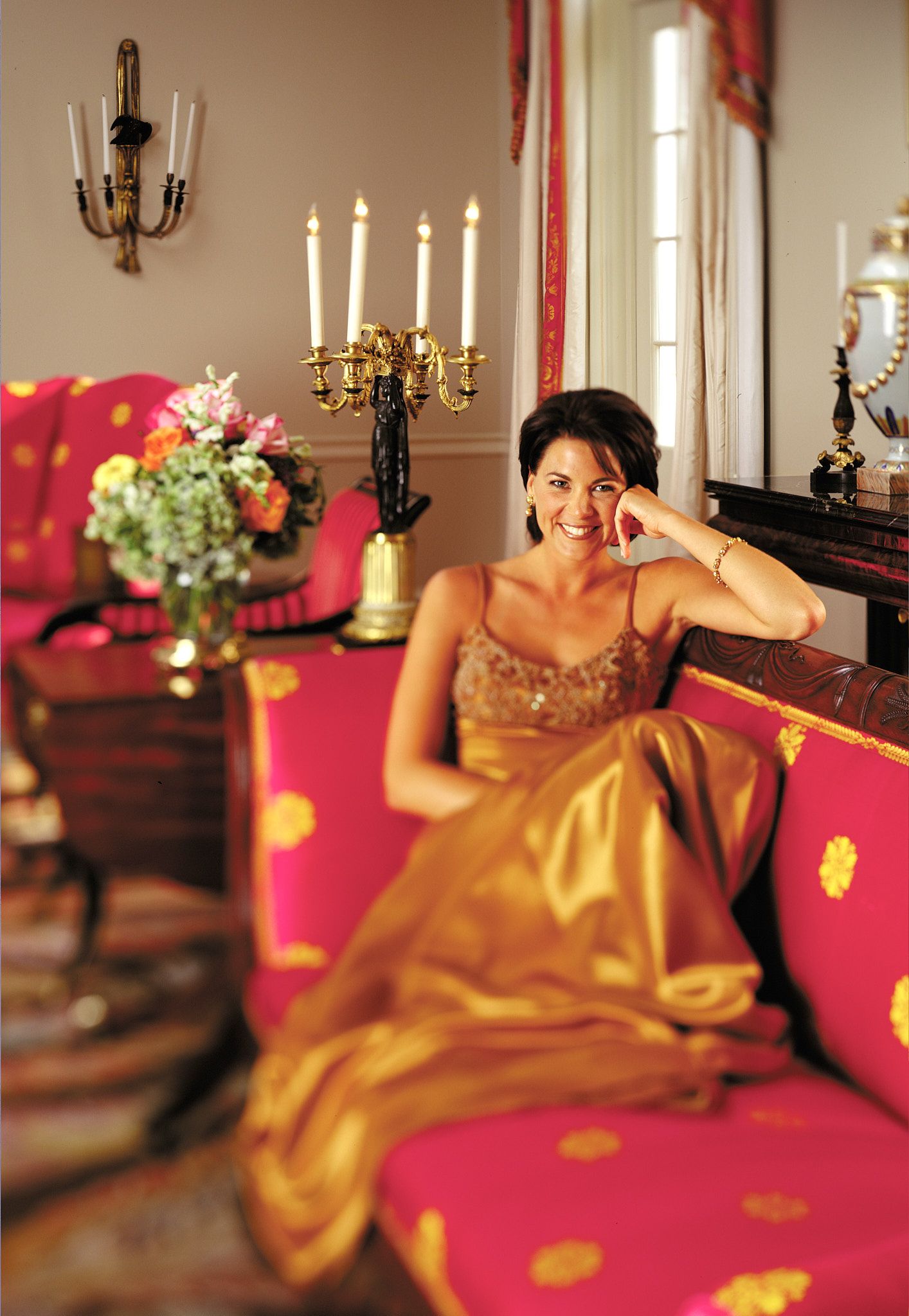 Smiling woman in a gold satin gown rests her chin on her hand on a pink sofa with gold floral pattern beside a candelabra and flowers.