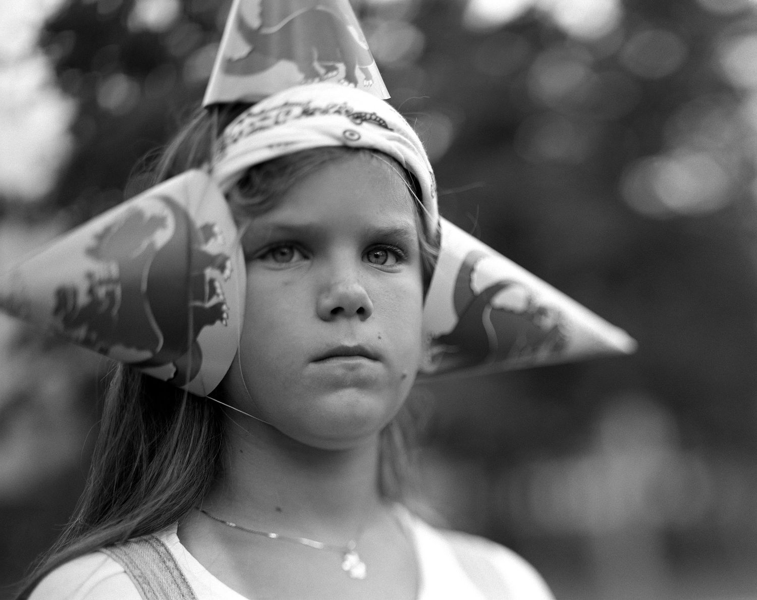 Black and white portrait of a serious young girl wearing a patterned party hat and a necklace outdoors.