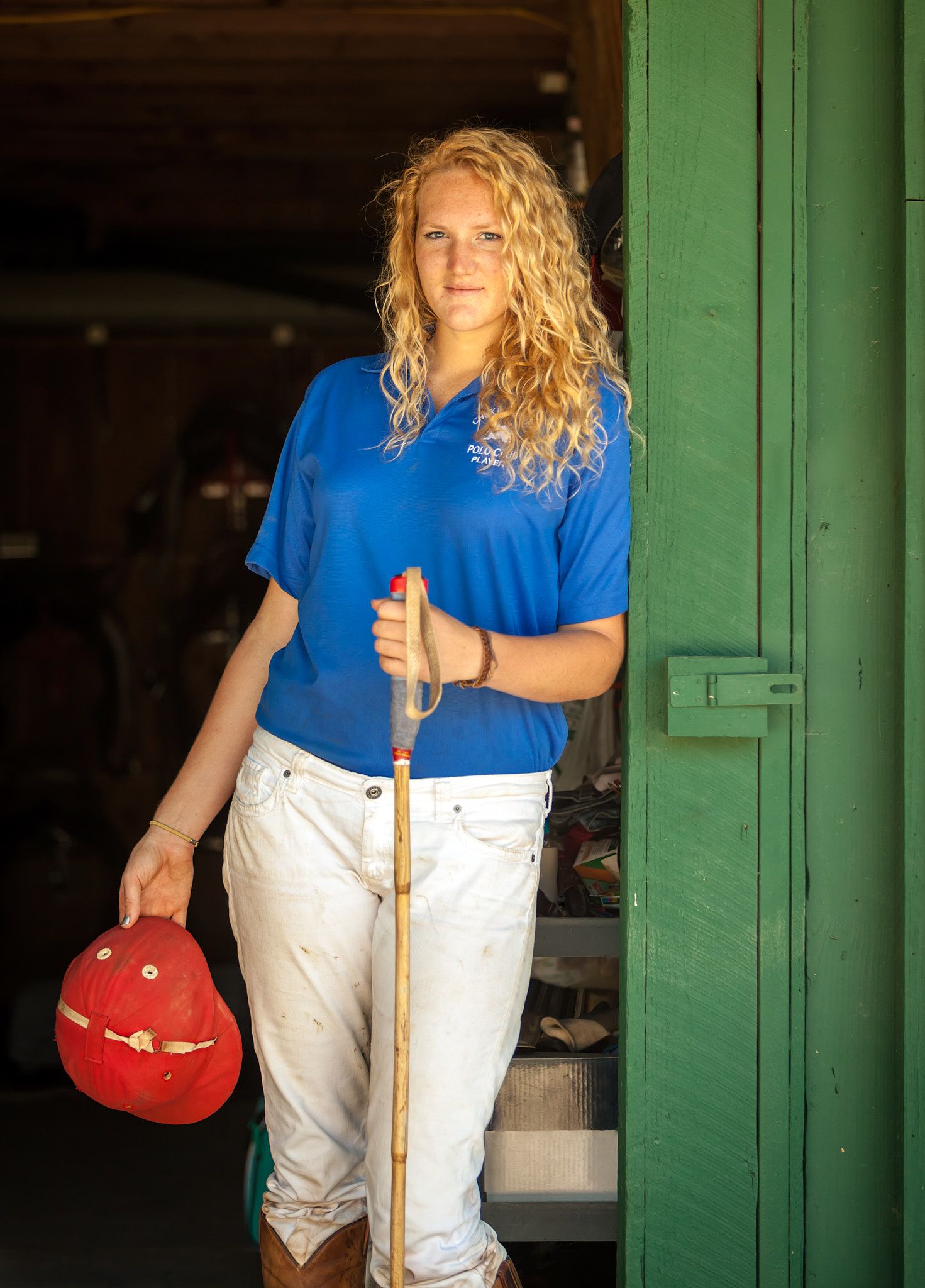 Young woman with curly blonde hair, blue polo and white breeches, holding a red riding helmet and wooden crop in a green stable doorway.