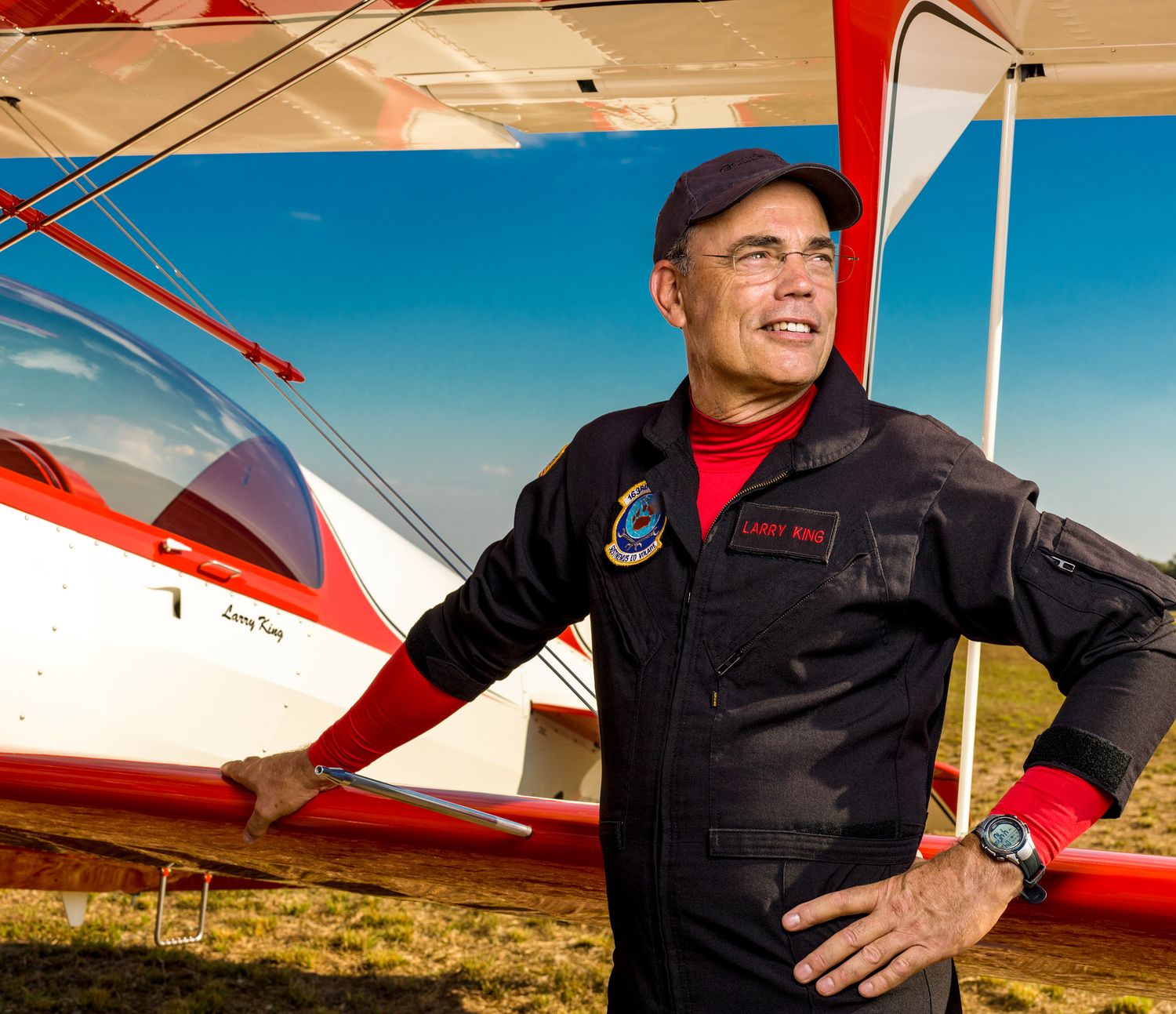 Smiling pilot in a navy flight suit and cap stands beside a red and white biplane on a grassy airfield under a bright blue sky.