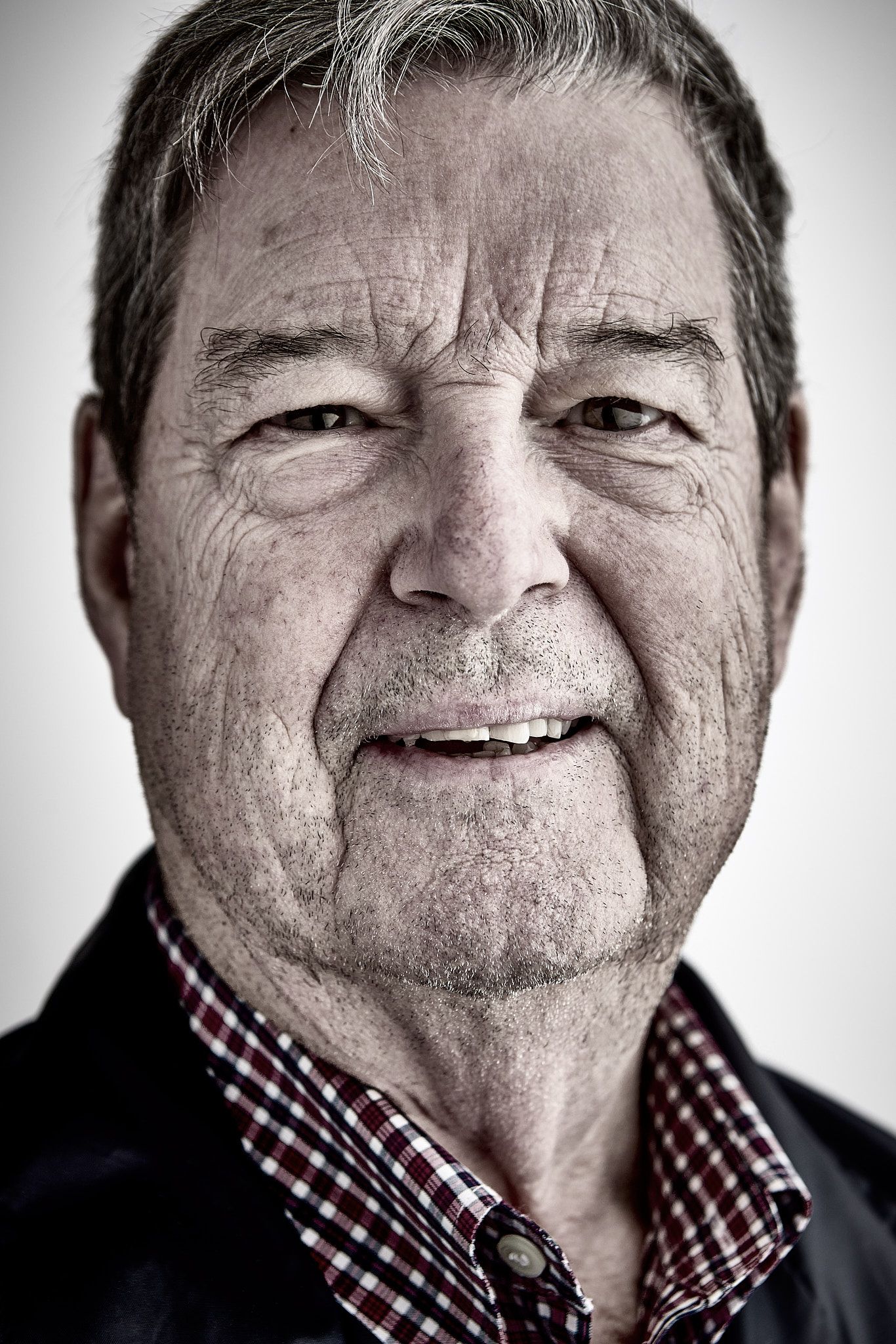 Close-up black and white style portrait of a smiling older man with gray hair wearing a checkered collared shirt and dark jacket.