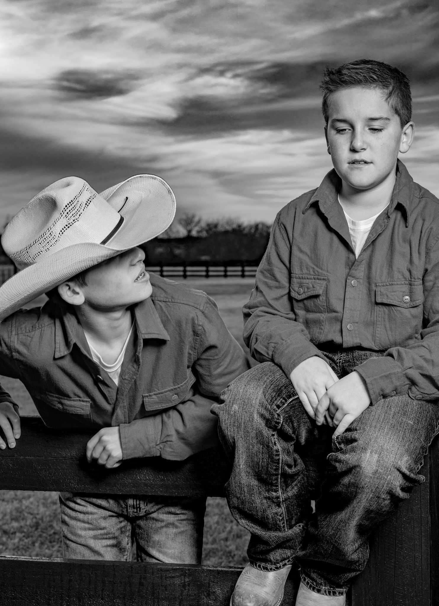 Black and white photo of two boys on a wooden ranch fence, one wearing a cowboy hat, under a dramatic cloudy sky.