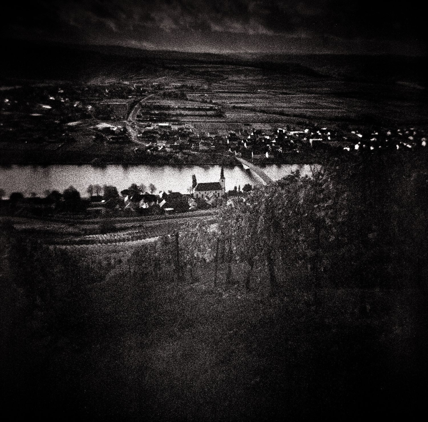 Moody black and white photo of a vineyard hillside overlooking a river, a church, and a distant village under a dramatic, cloudy sky.