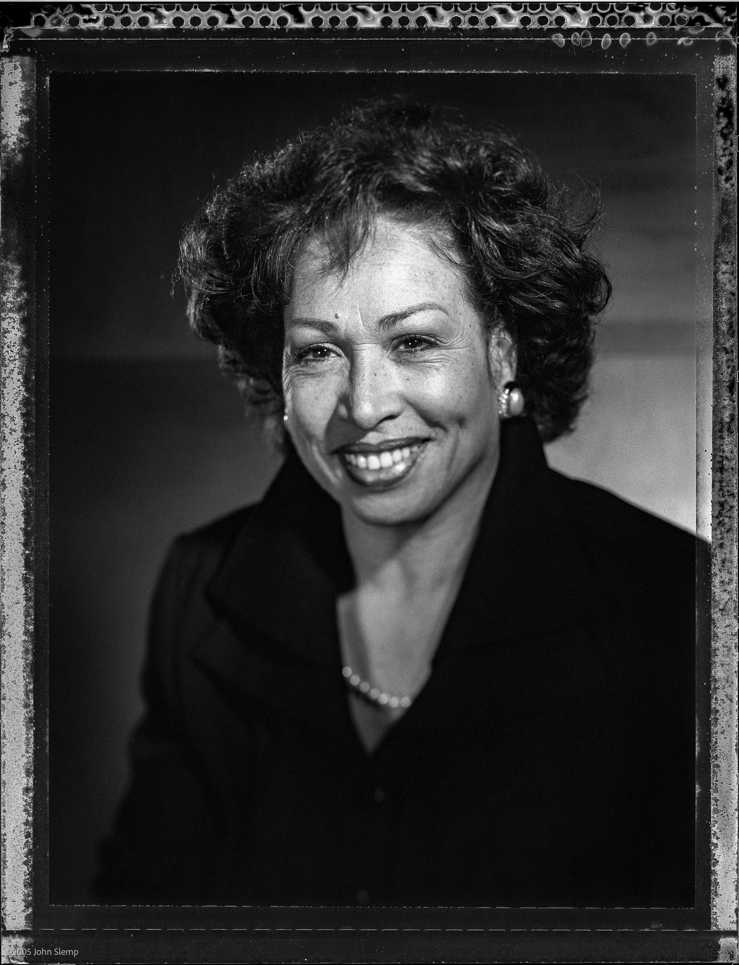 Black and white studio portrait of a smiling woman with curly hair wearing a dark jacket and pearl necklace.