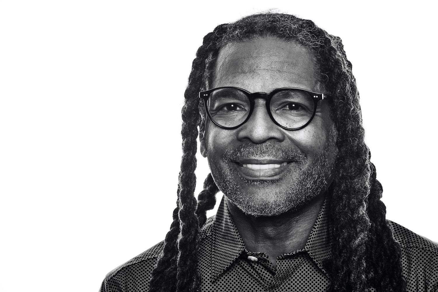 Black and white portrait of a smiling man with long locs, round dark-framed glasses, and a patterned button-up shirt.