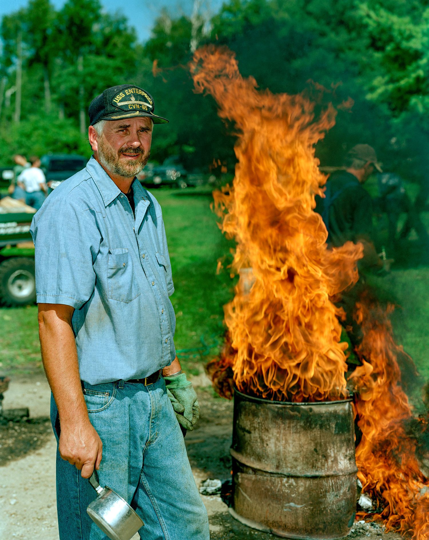 Man in a blue work shirt, jeans and cap holds a ladle beside a metal barrel with tall orange flames rising outdoors on a sunny day.