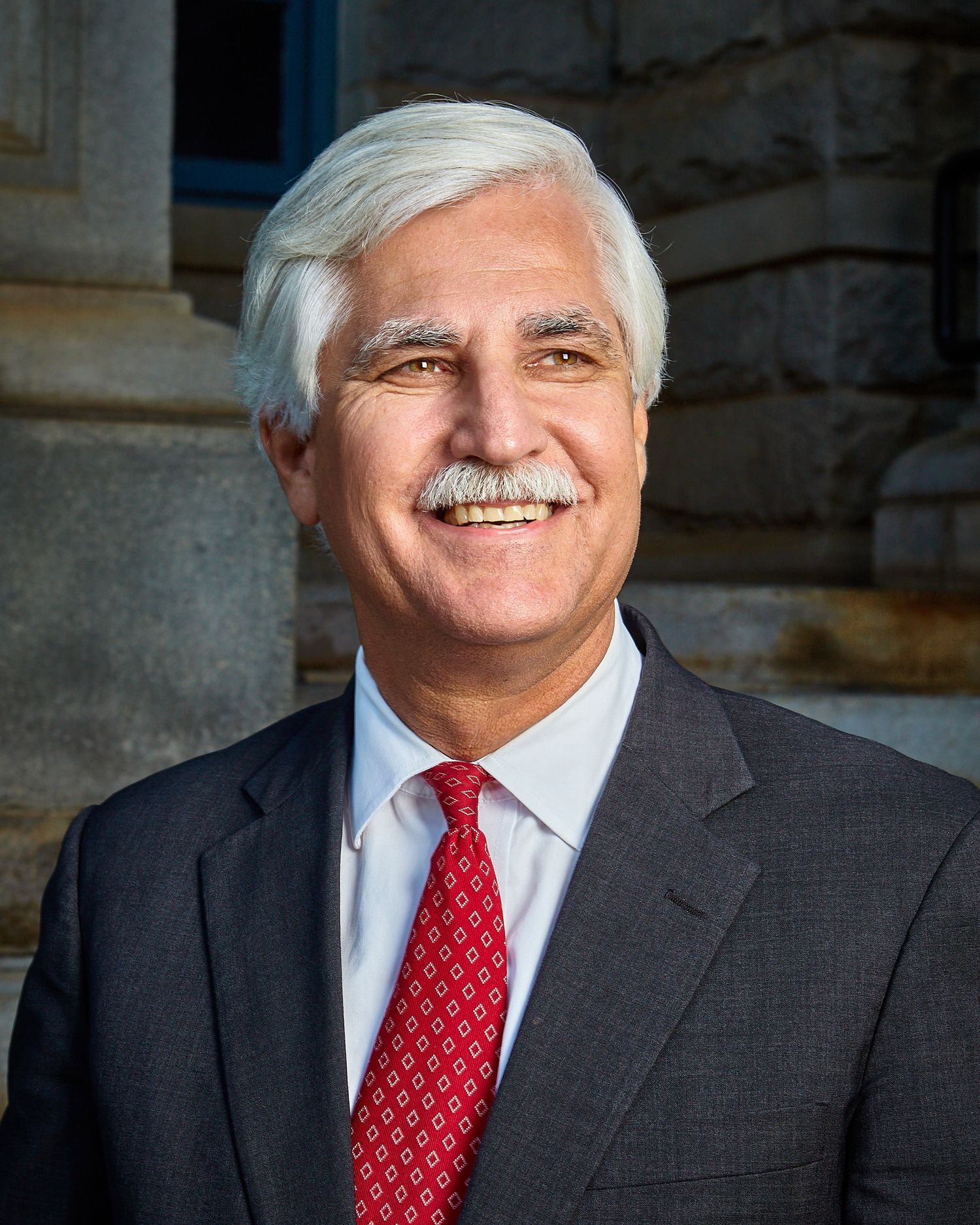 Smiling man with white hair and a mustache wearing a gray suit, white shirt and red patterned tie in front of a stone building.