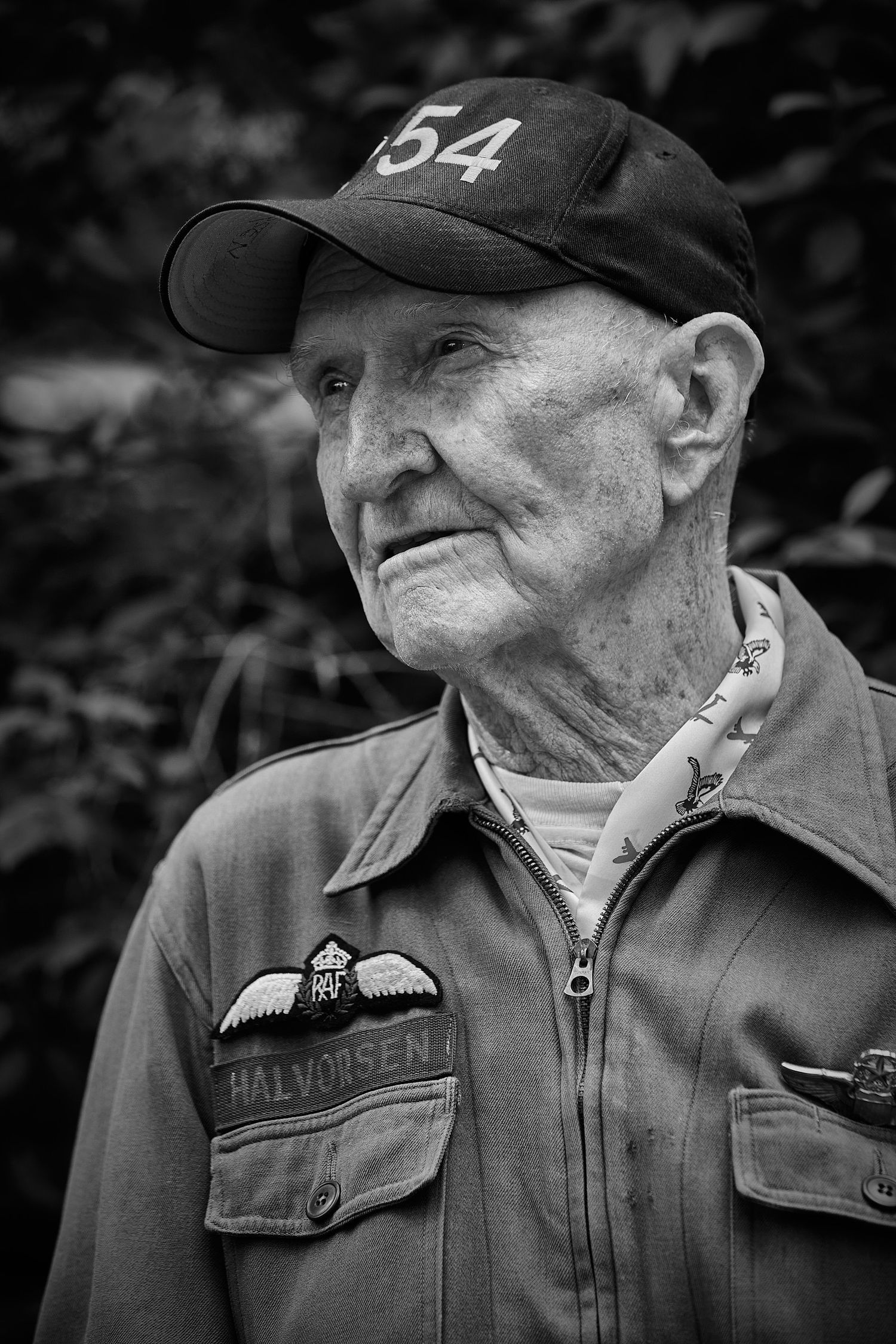 Black and white portrait of an elderly man in a cap numbered 54 and a work jacket with a winged name patch, gazing off to the side.