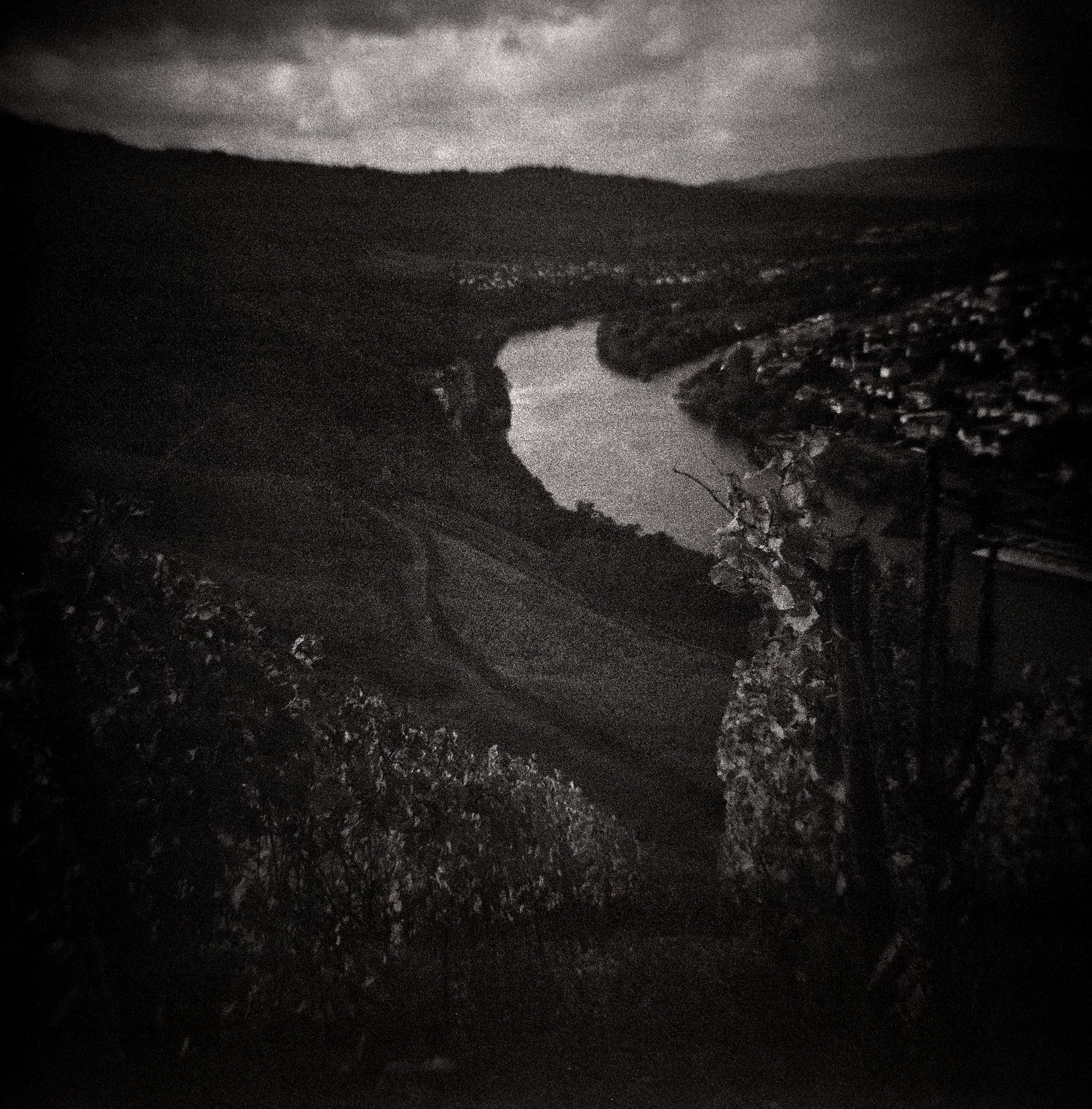 Moody black and white landscape of a winding river curving through a hillside valley with vineyards, a distant town and a stormy sky.
