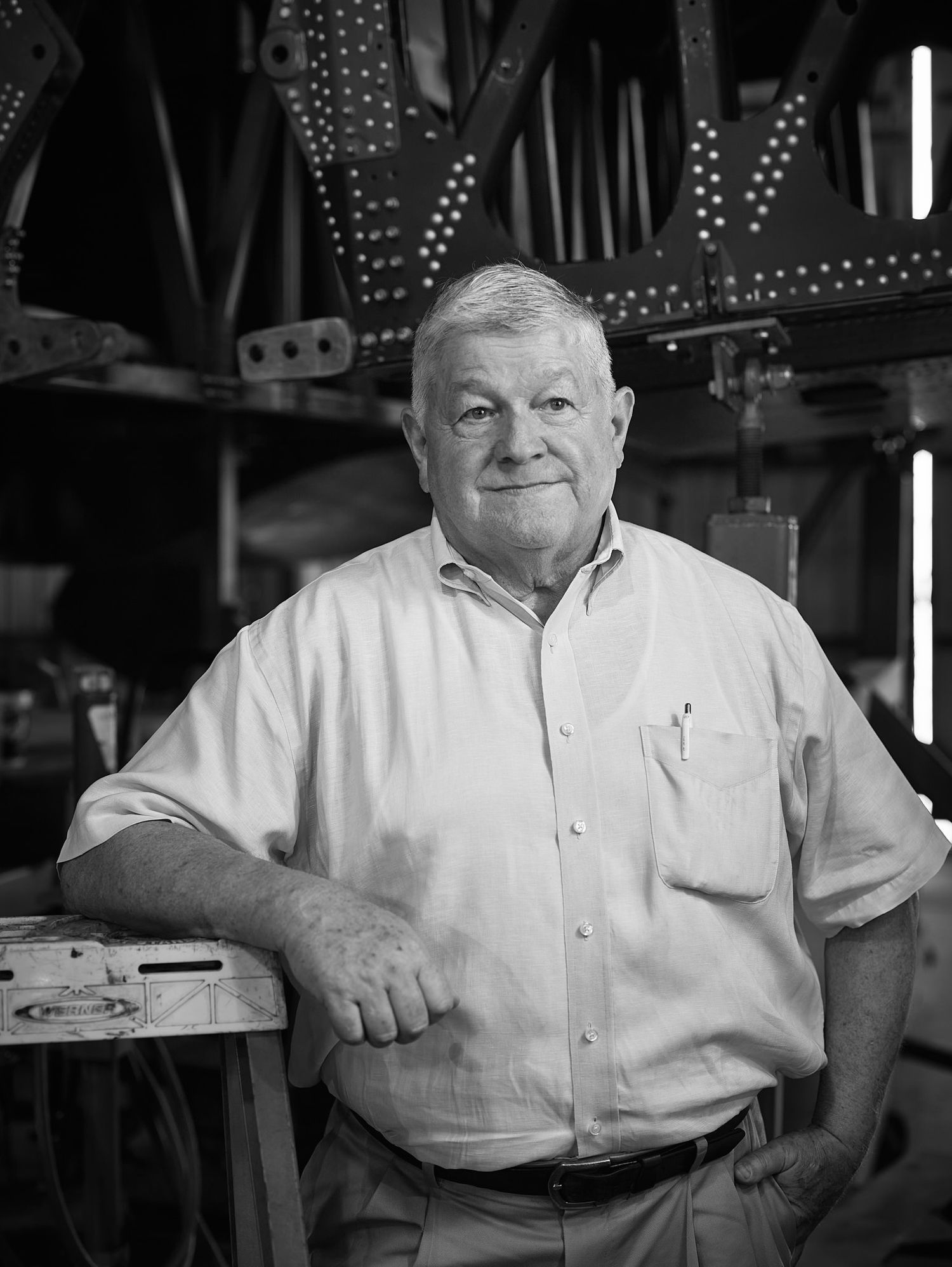 Black and white portrait of a smiling older man in a button-up shirt leaning on a workbench in an aircraft hangar.