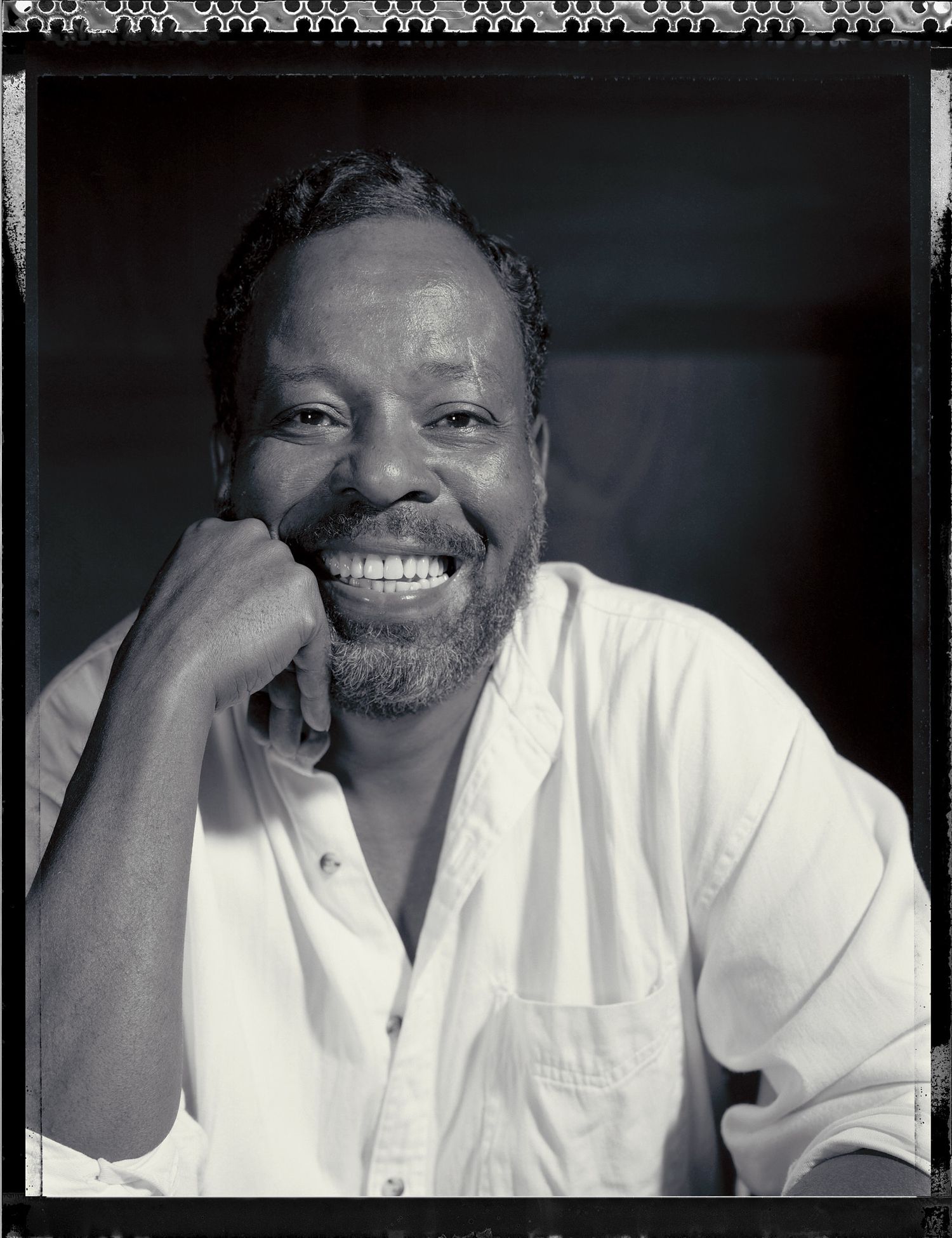 Black and white portrait of a smiling bearded man in a white shirt resting his chin on his fist.