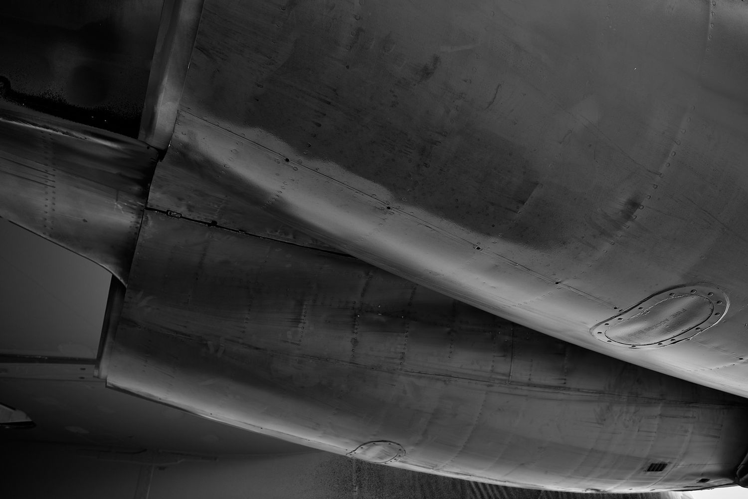 Black and white detail of an aircraft engine nacelle and fuselage underside showing weathered metal panels and rivets.