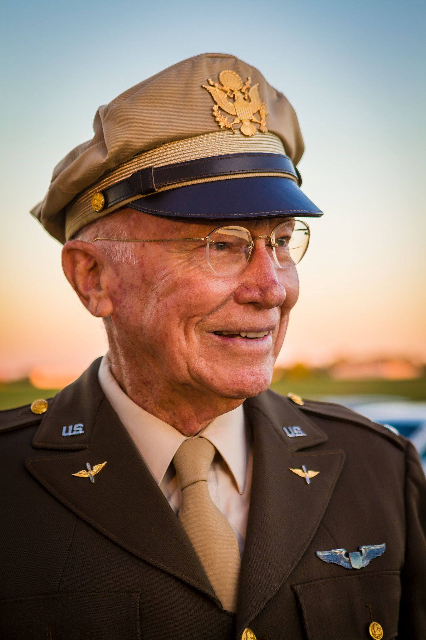 Smiling elderly man in a vintage U.S. military dress uniform with eagle-insignia cap, wings pin, and glasses, photographed at sunset.