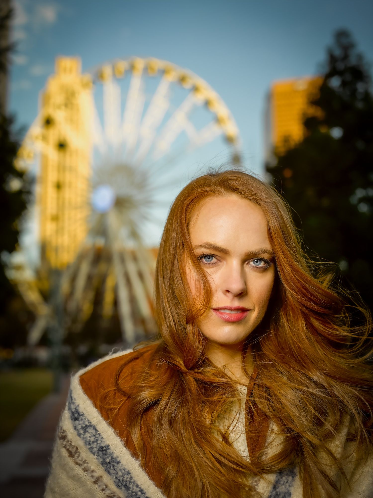 Young woman with long auburn hair and blue eyes in a patterned wool wrap, posing before a lit Ferris wheel at golden hour.