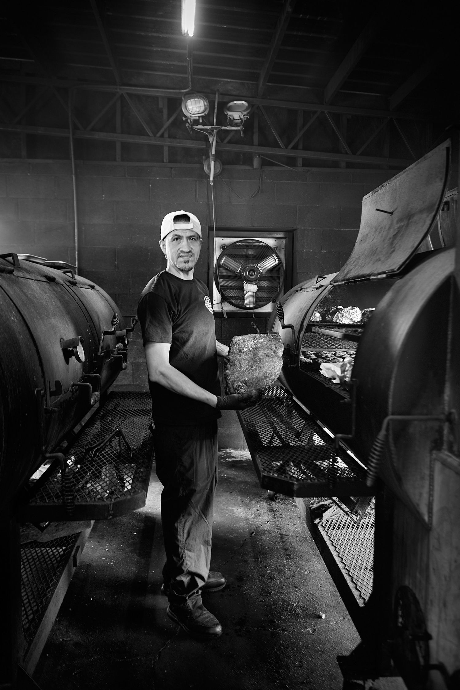 Black and white photo of a man in a white cap holding a large piece of meat beside industrial barbecue smokers in a dim smokehouse.