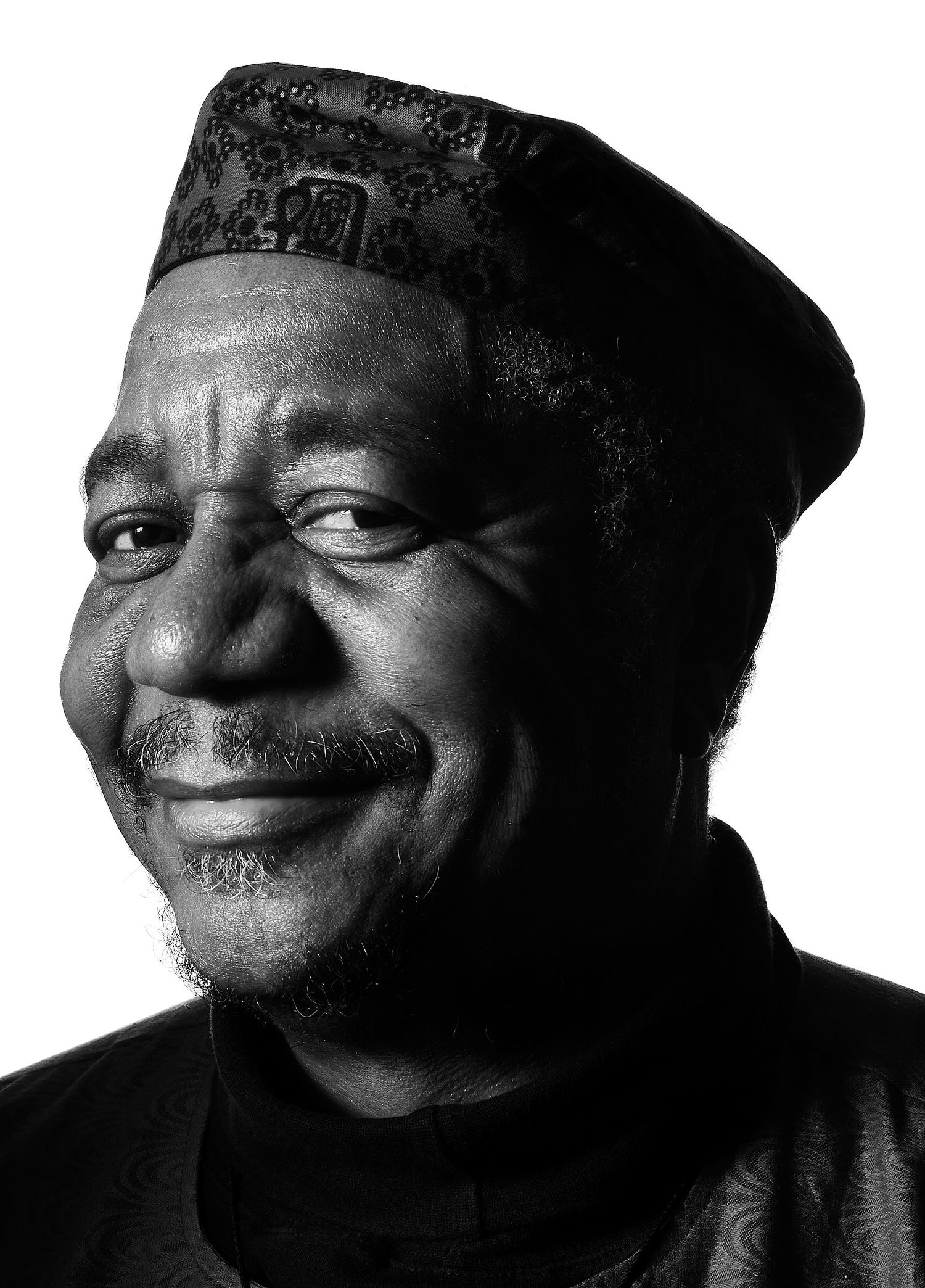Black and white close-up portrait of a smiling older man with a goatee wearing a patterned embroidered cap and a high-collared top.