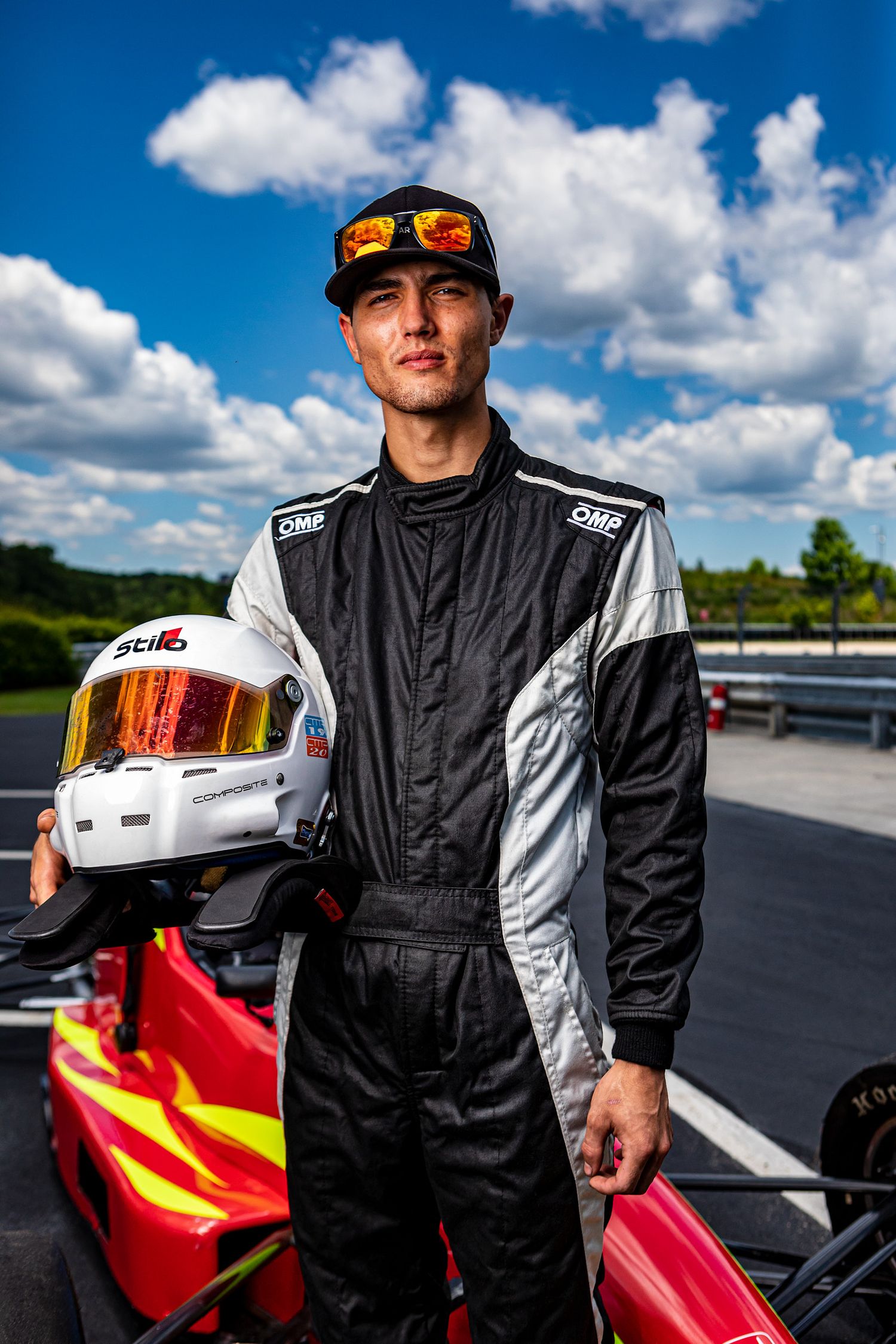 Race driver in black and white OMP racing suit and cap holding a white Stilo helmet beside a red go-kart on a sunny track.