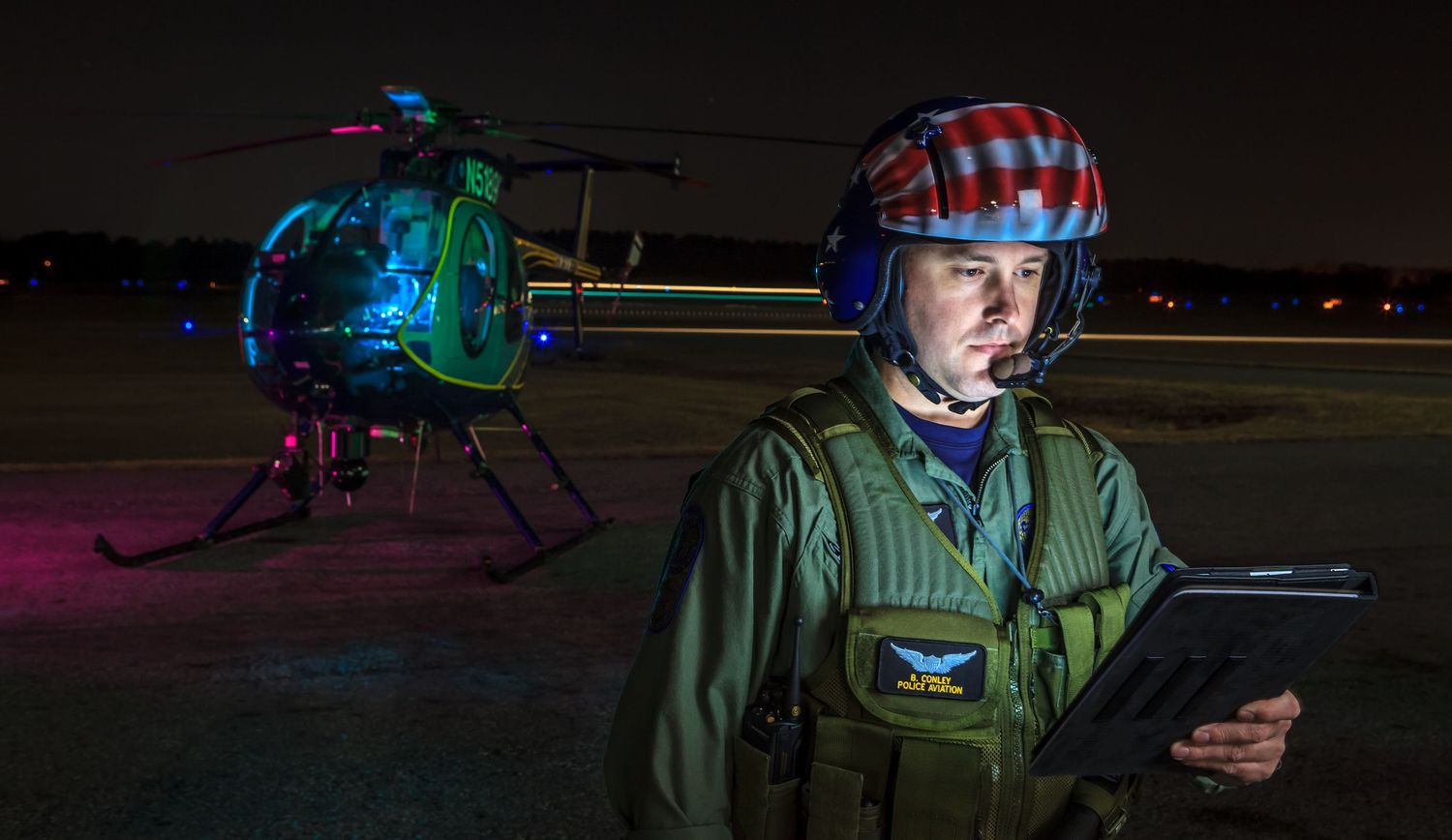 Helicopter pilot in a flight helmet and green vest checks a tablet on a night airfield beside an illuminated helicopter.