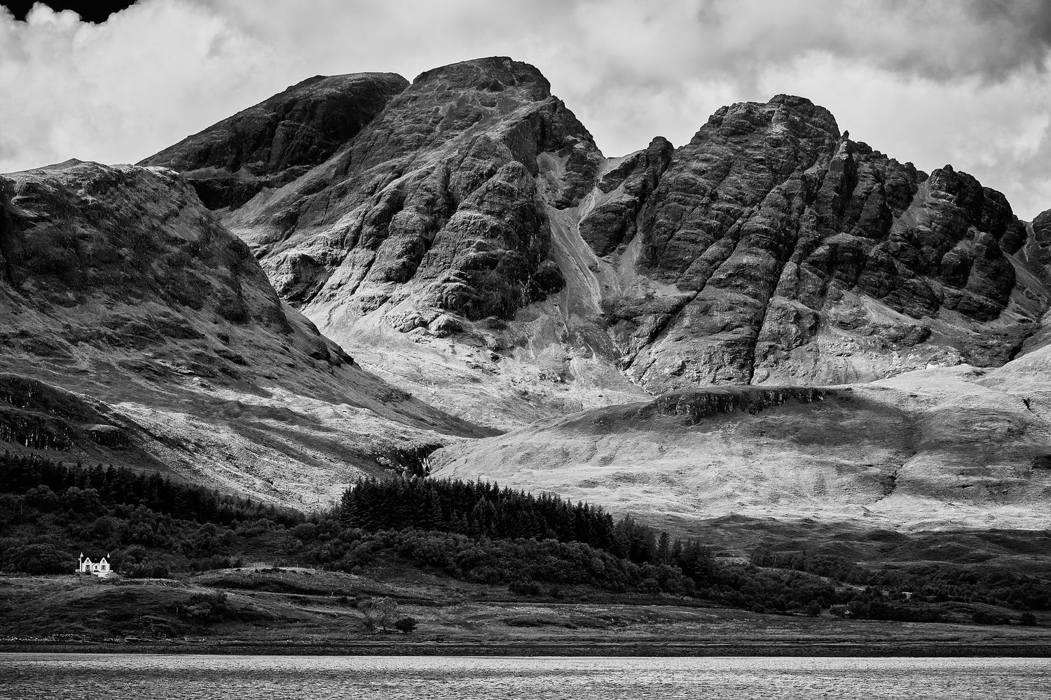 Black and white photograph of craggy mountains above a quiet sea loch, with a lone white cottage on the shoreline below.