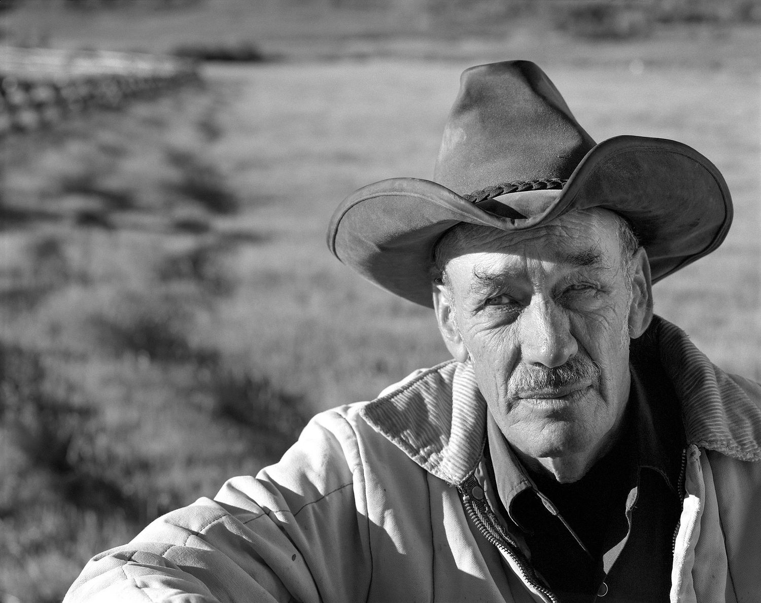 Black and white portrait of a weathered older man in a wide-brimmed cowboy hat and jacket standing in an open field.