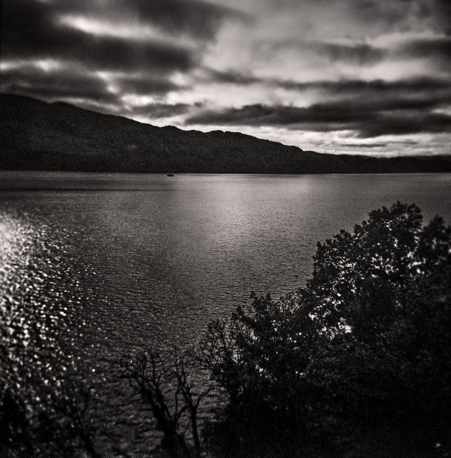 Black and white landscape of a calm lake beneath dramatic storm clouds, with a dark mountain ridge and silhouetted trees in the foreground.