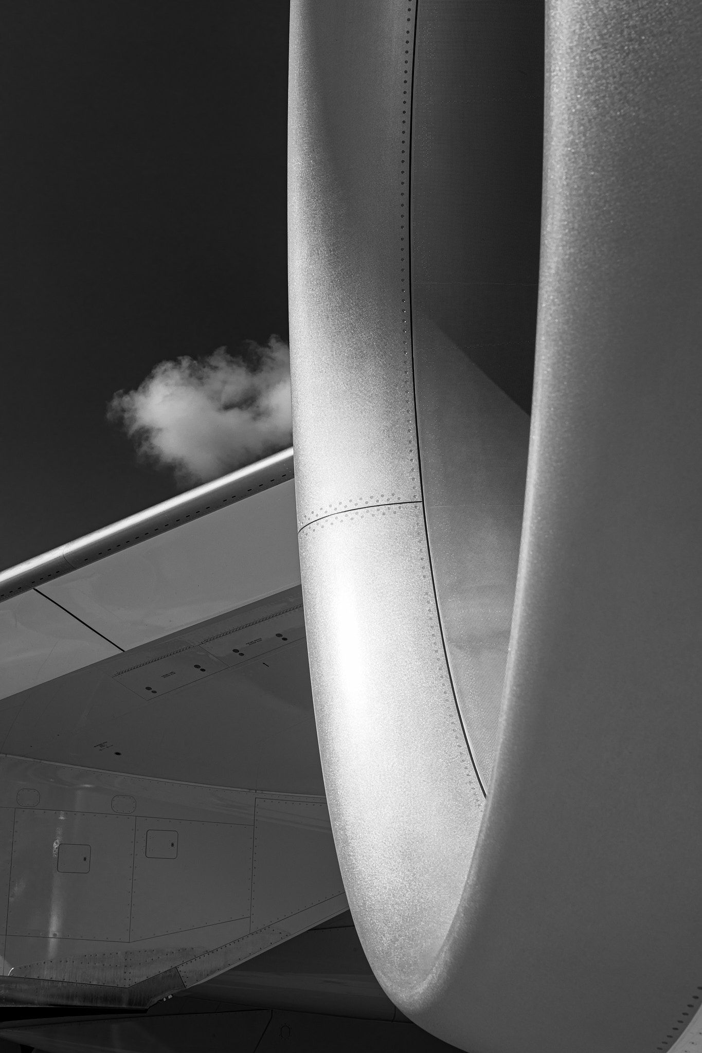 Black and white close-up of a jet engine nacelle curving beside an aircraft wing with a small cloud in the dark sky.