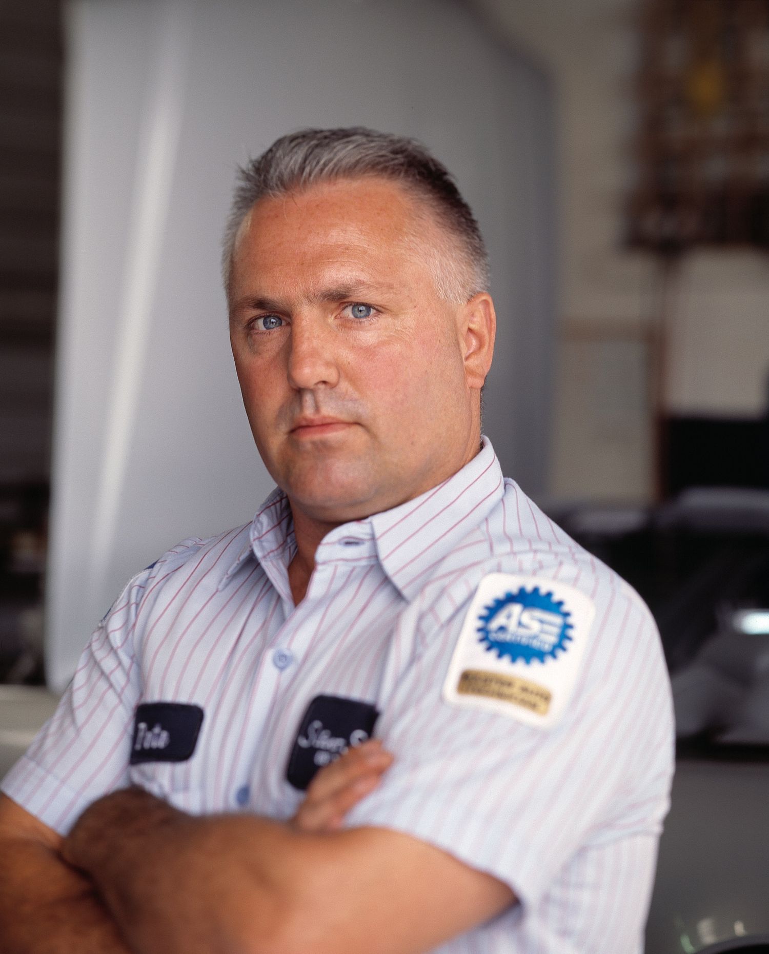 Auto mechanic with gray hair and crossed arms wears a striped work shirt with an ASE certification patch inside a repair shop.