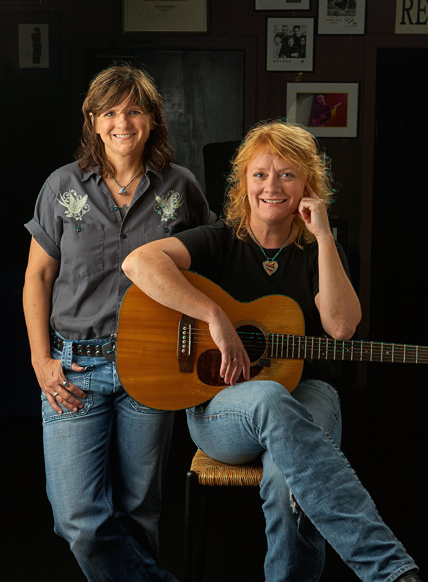 Two smiling women pose in a dimly lit room, one standing in a gray embroidered shirt and the other seated holding an acoustic guitar.