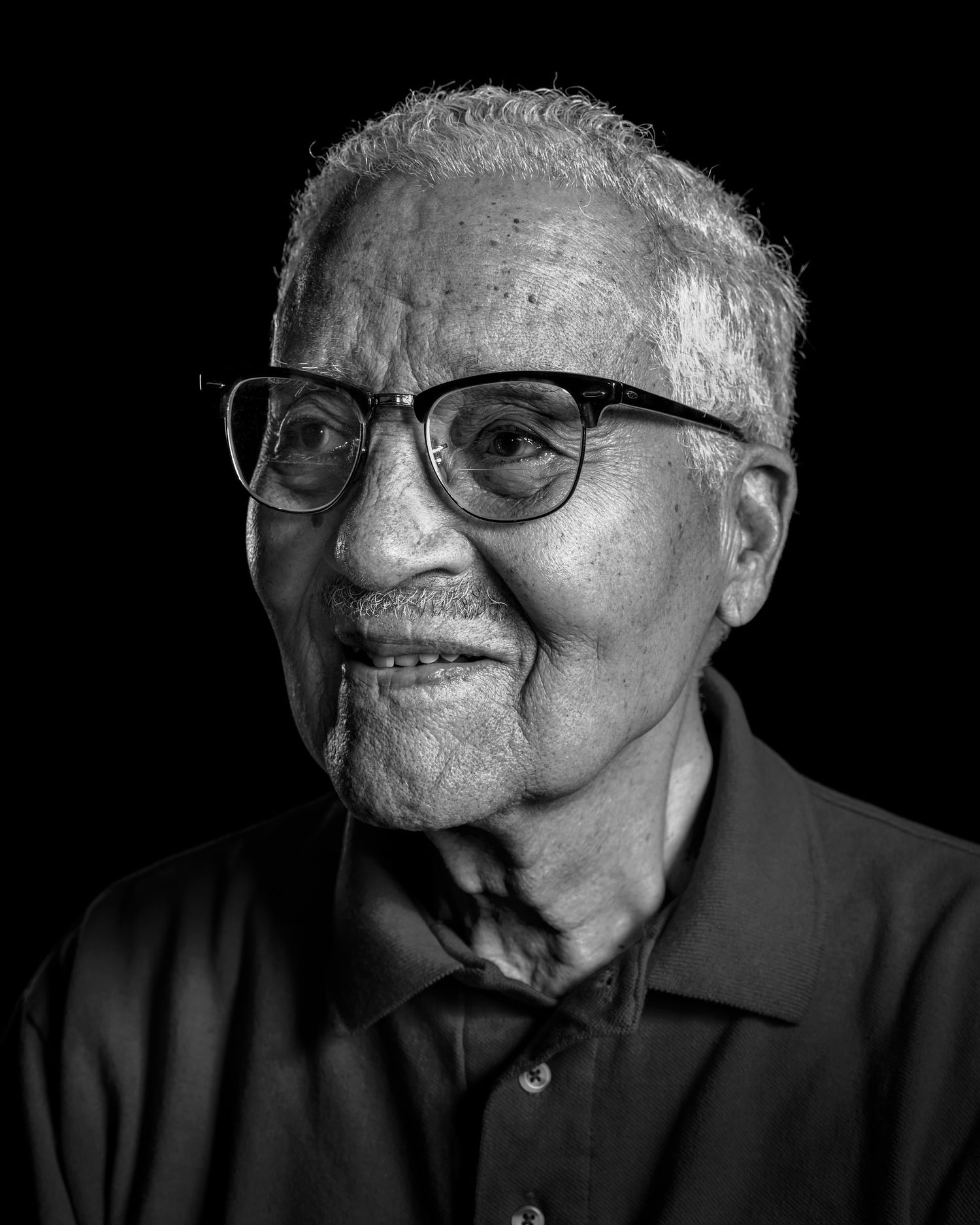 Black and white studio portrait of a smiling elderly man with white hair and dark-rimmed glasses wearing a collared polo shirt.